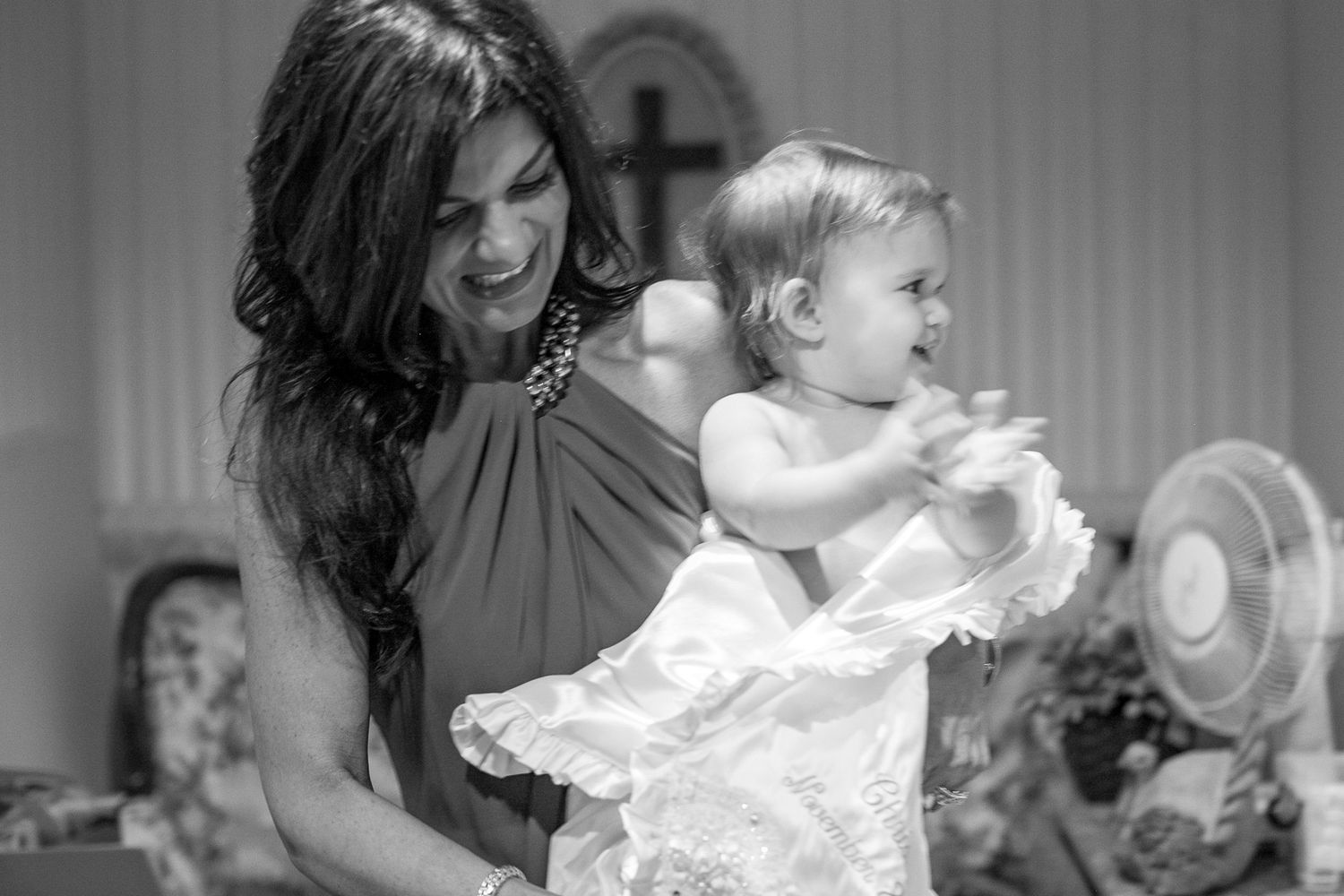 Black and white photo of a woman holding a baby in a white christening gown, smiling in a church setting.