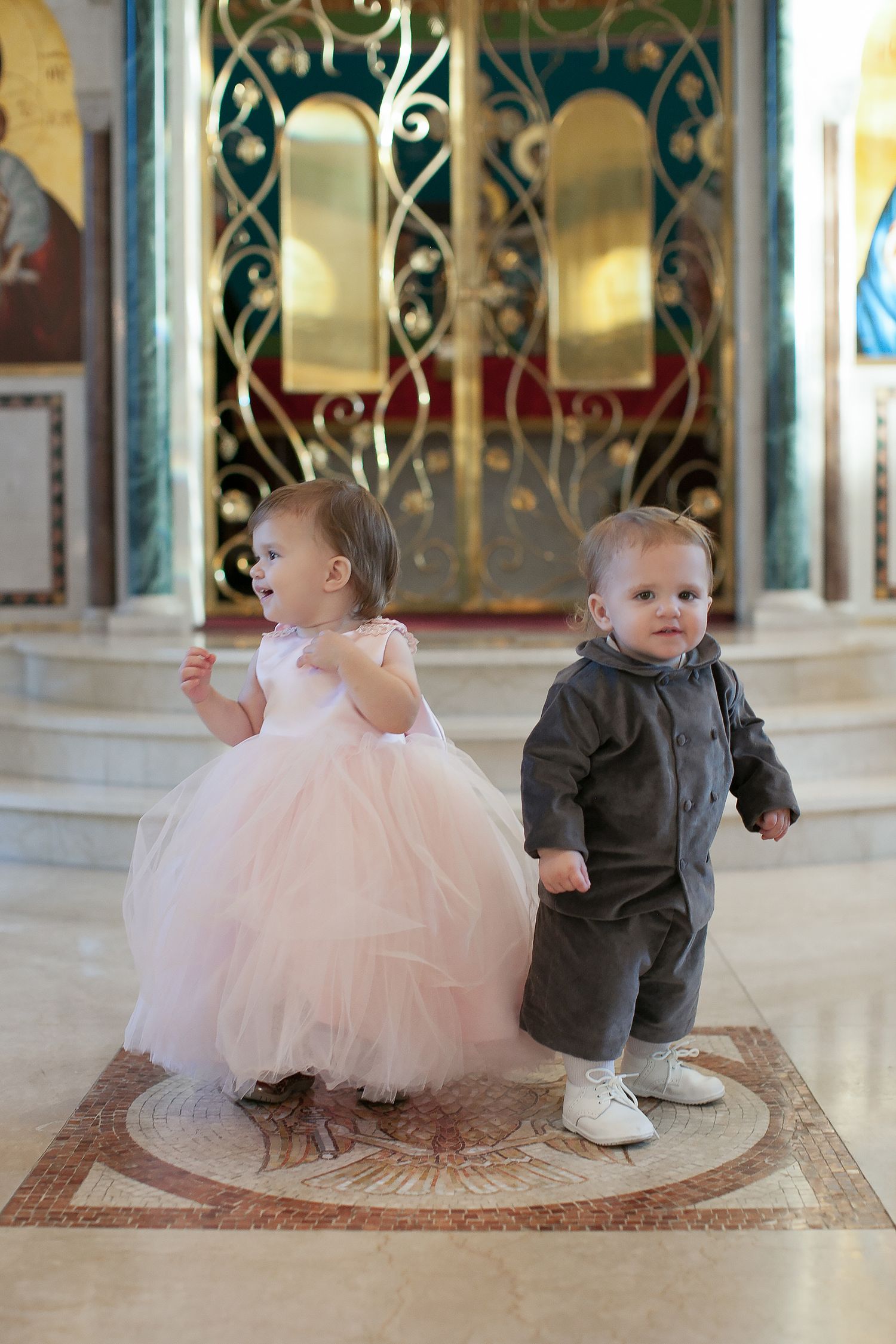 Two toddlers dressed formally stand in an ornate church, a girl in a pink tulle dress and a boy in a gray suit.