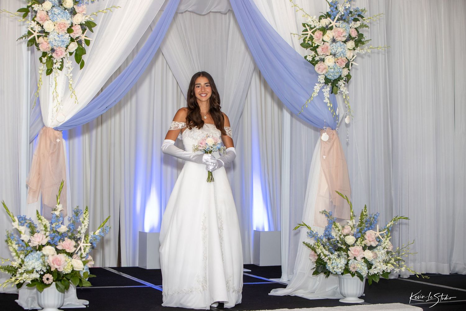 A smiling bride in a white gown and gloves holds a bouquet under a floral arch with blue and pink flowers.