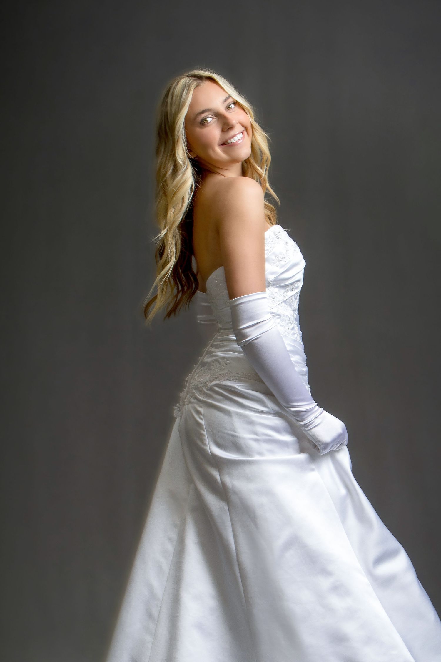 Smiling blonde bride in white strapless satin gown and long white gloves poses elegantly against gray background.