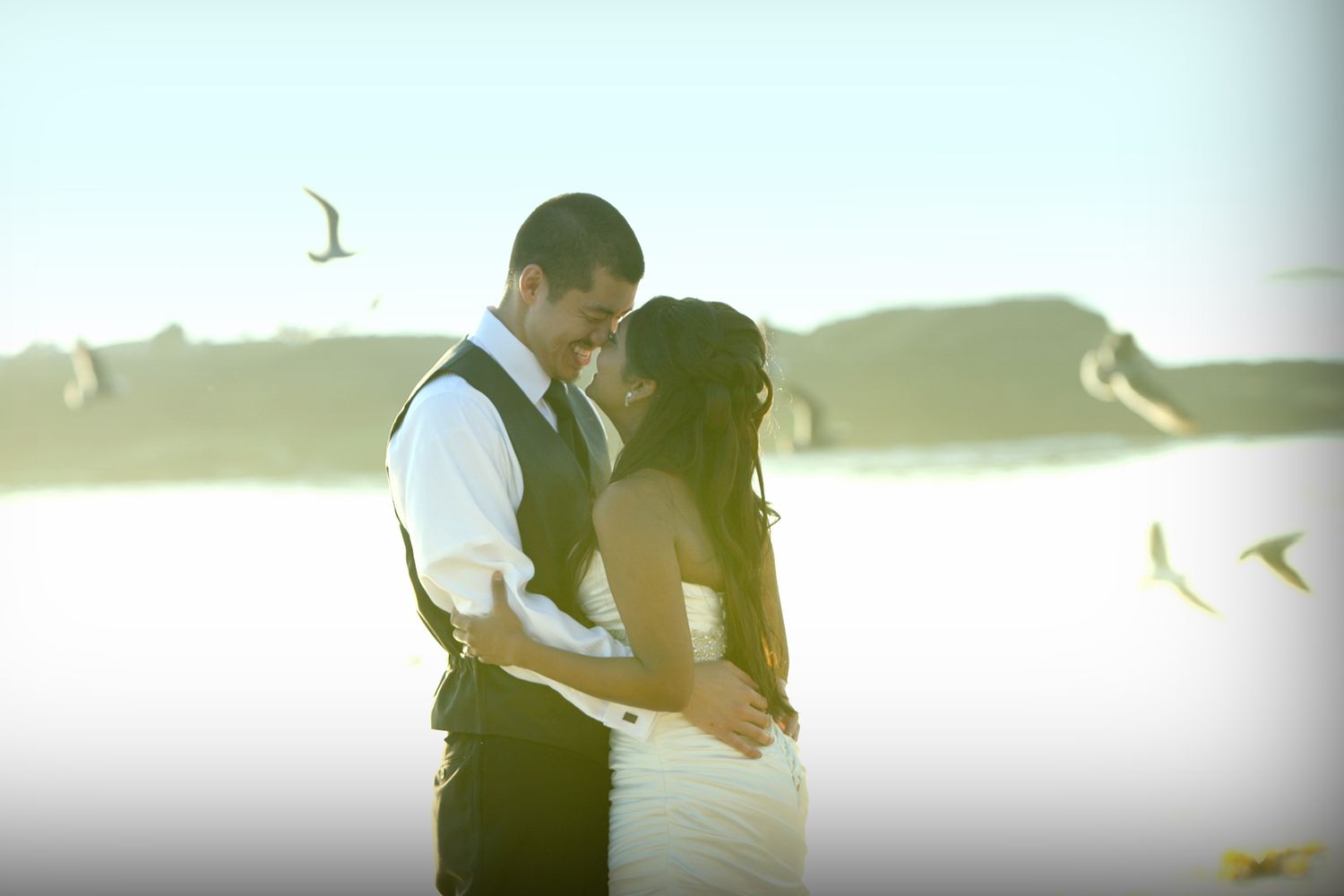 Romantic couple sharing an intimate moment on a sunlit beach with birds flying in the background.