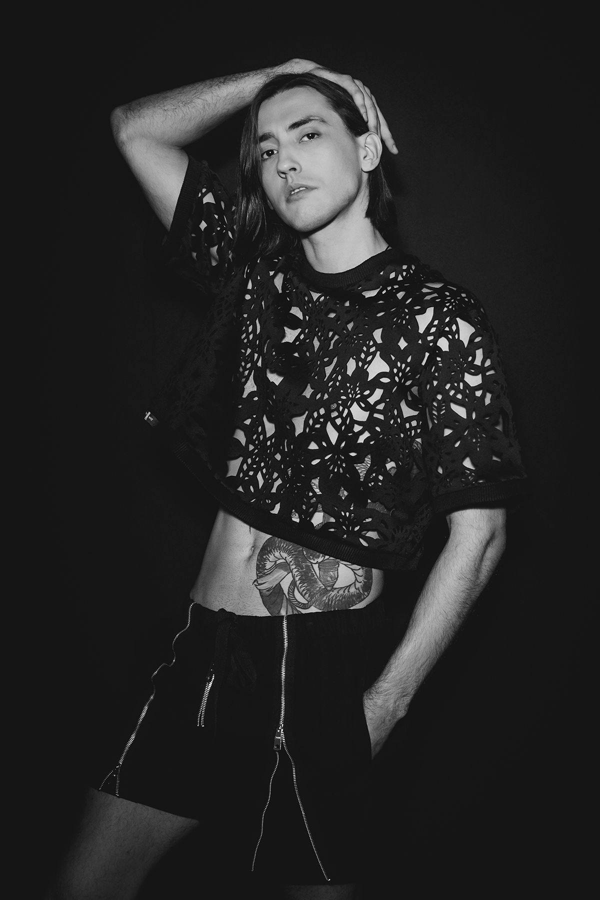 Black and white portrait of a young man in lace crop top with tattoo, posing confidently against dark background.