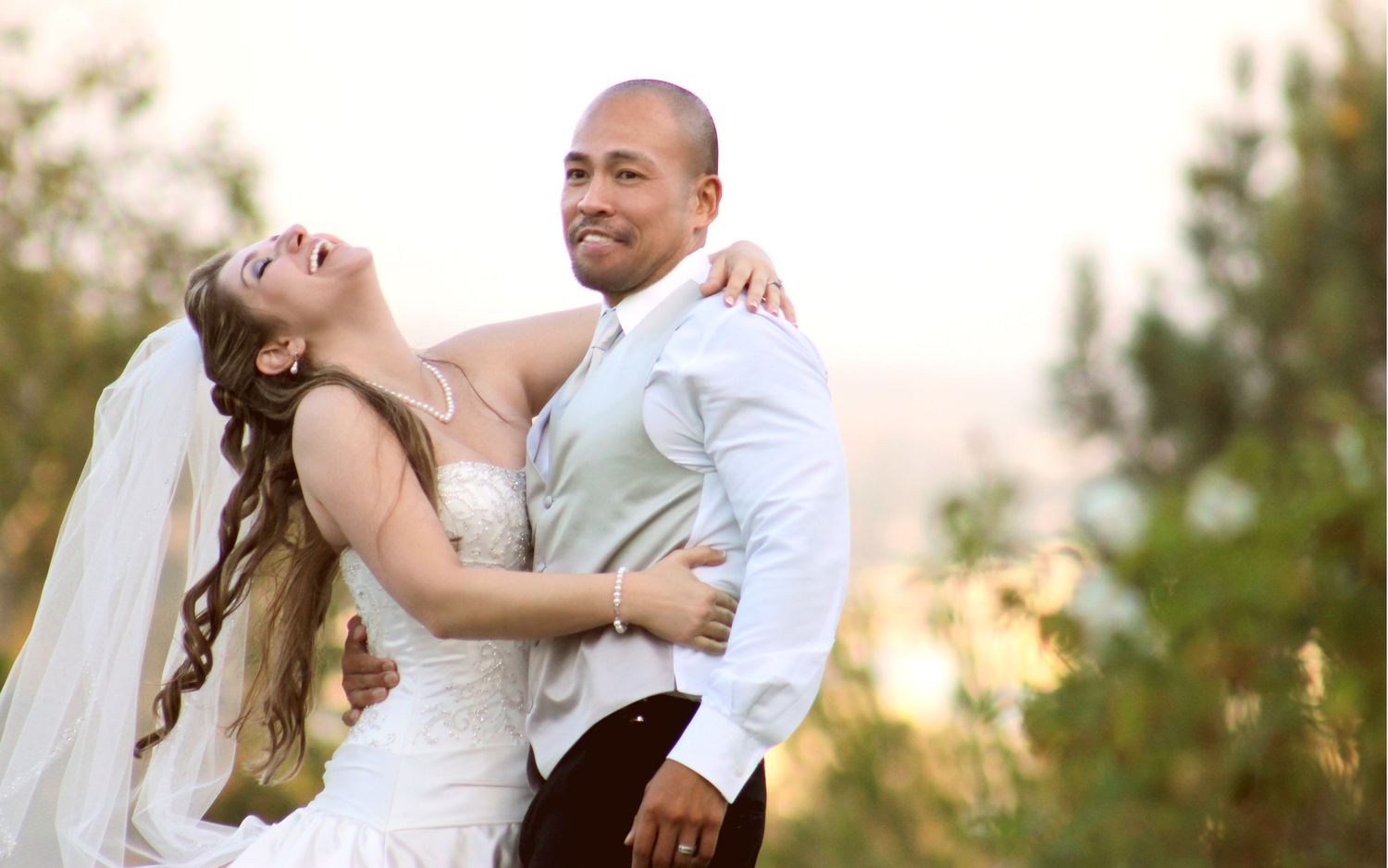 A bride and groom share a joyful embrace outdoors, with the bride in a white gown and veil on their wedding day.