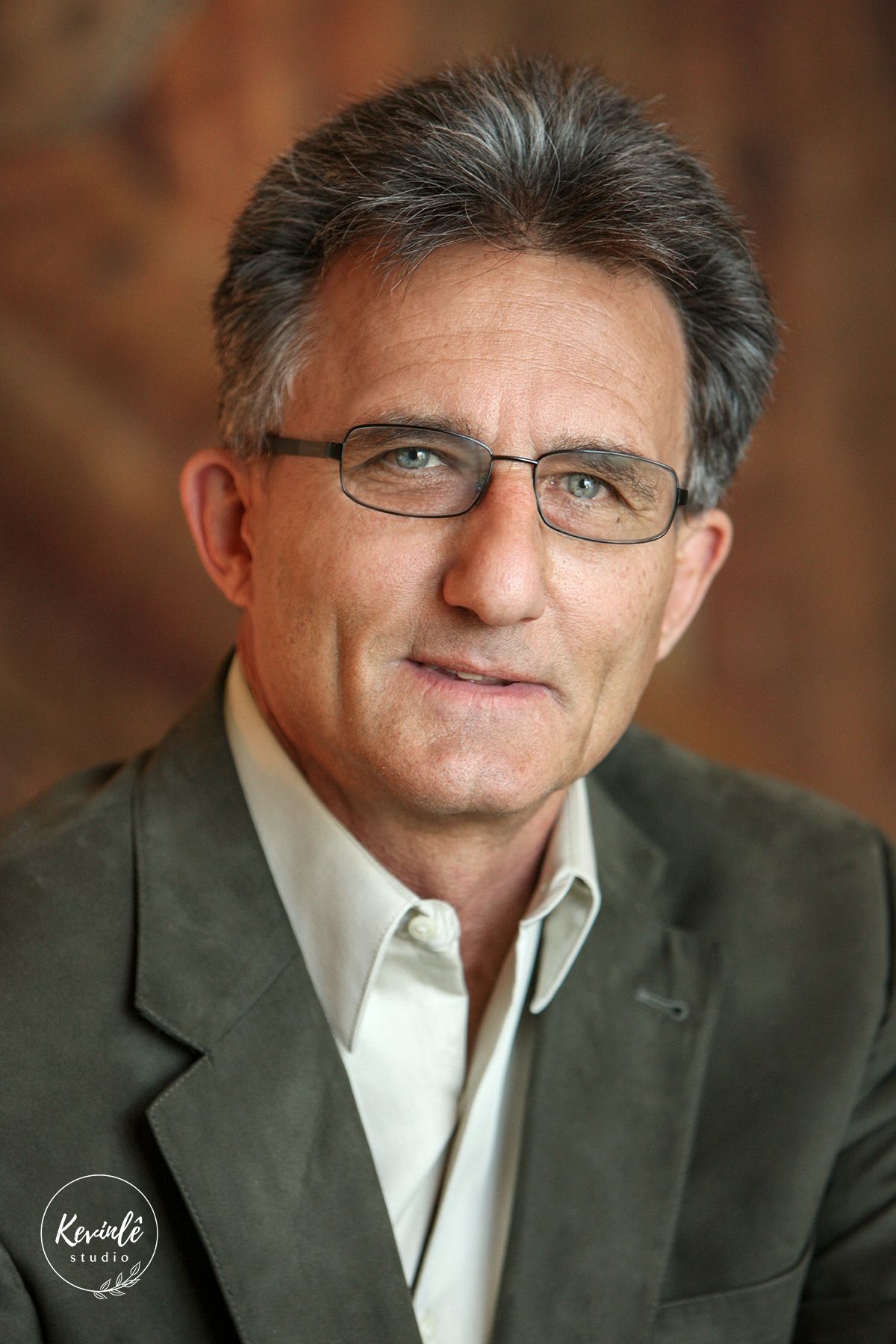 Middle-aged man with gray-streaked hair and glasses wearing a gray blazer, smiling confidently in a professional portrait.