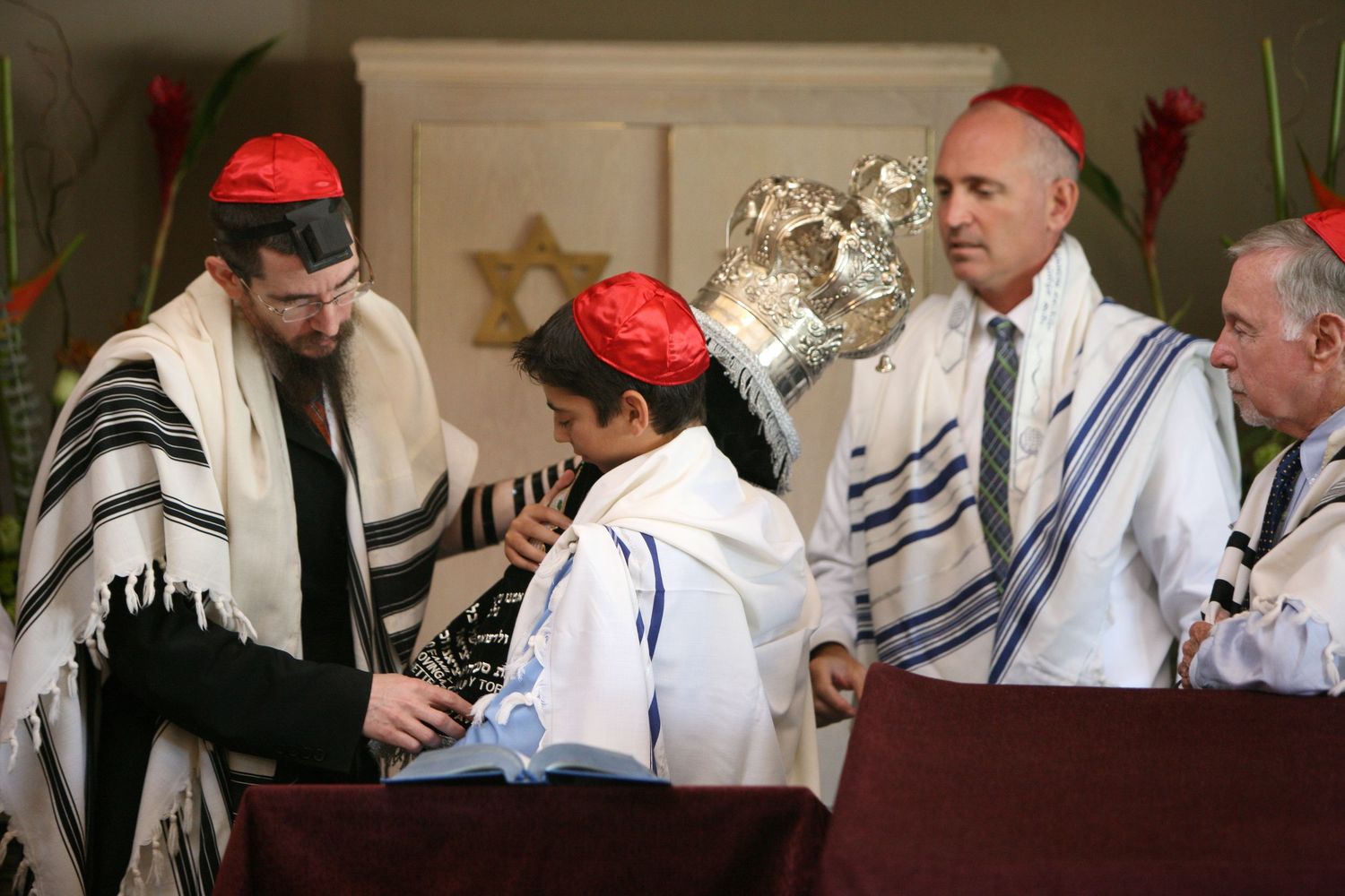 Jewish Bar Mitzvah ceremony with men wearing tallit prayer shawls and red kippot reading from the Torah.