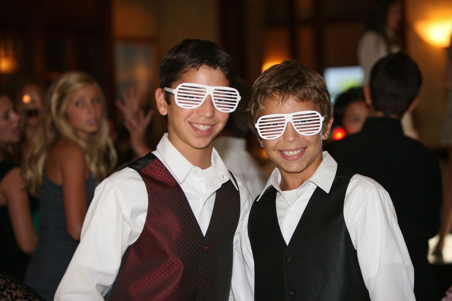 Two boys wearing matching white shutter shades and vests smile at a bar mitzvah party.