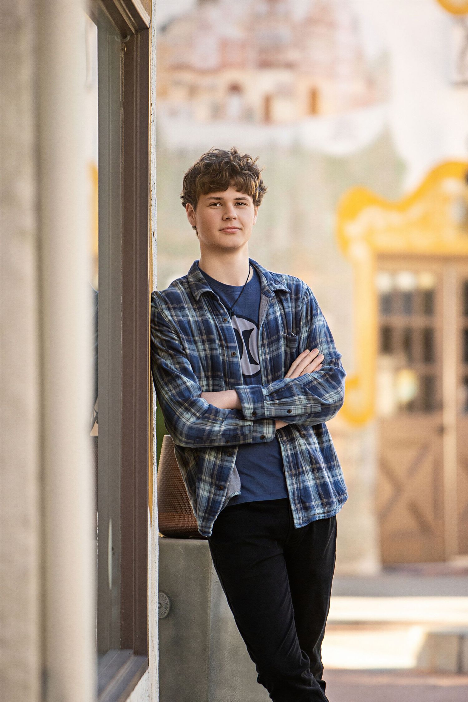 Teen boy in blue plaid shirt stands arms crossed against a wall outdoors in a urban setting.