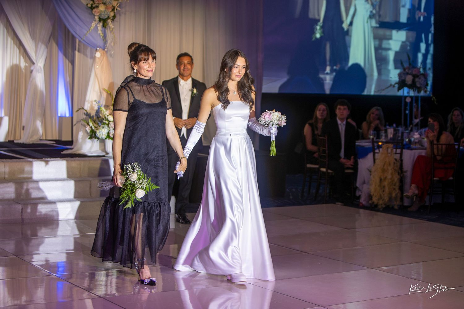 Bride in white gown walks with bridesmaid in black dress at elegant wedding reception venue.
