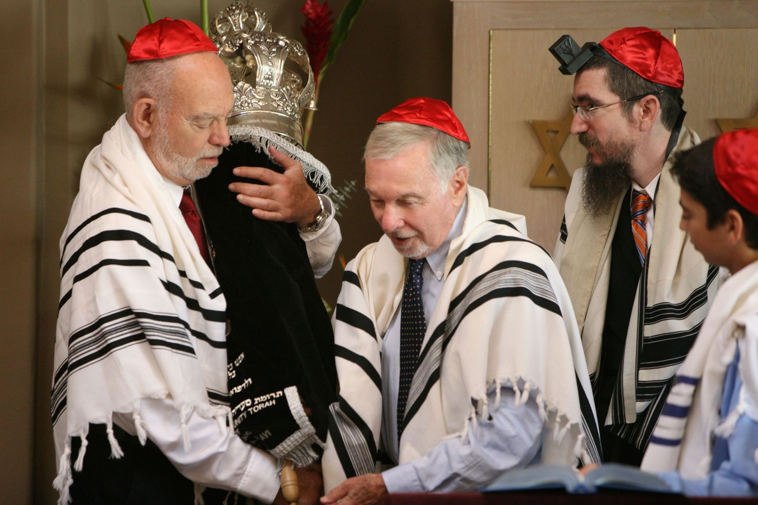 Jewish men in tallits and red kippot participate in a Torah ceremony during a synagogue service.