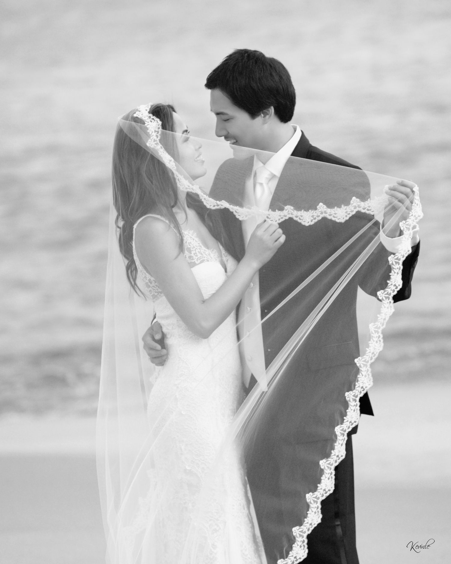 Black and white beach wedding photo of bride and groom sharing romantic moment, bride's lace veil flowing in the breeze.