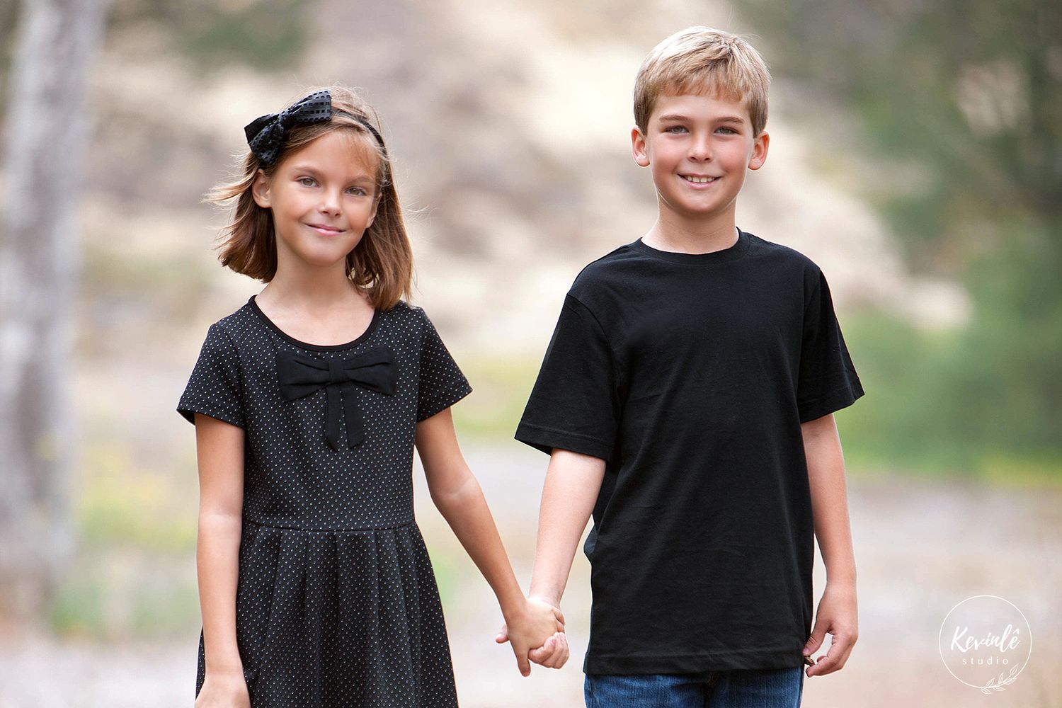A young girl in black dress and boy in black shirt hold hands outdoors in a professional portrait.