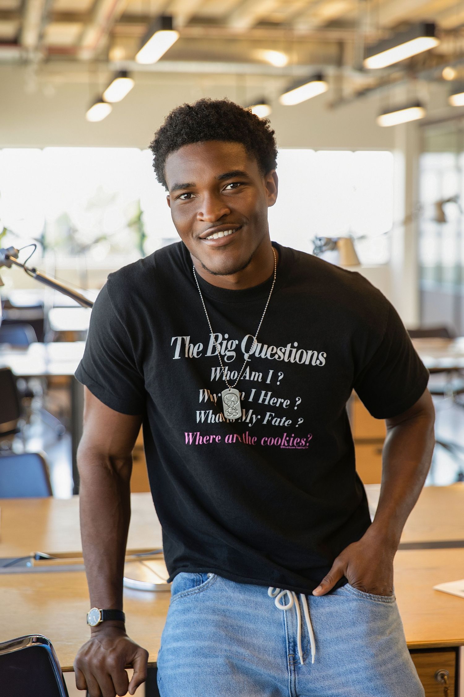 Smiling Black man in black graphic tee with Big Questions print and jeans, standing in modern office.