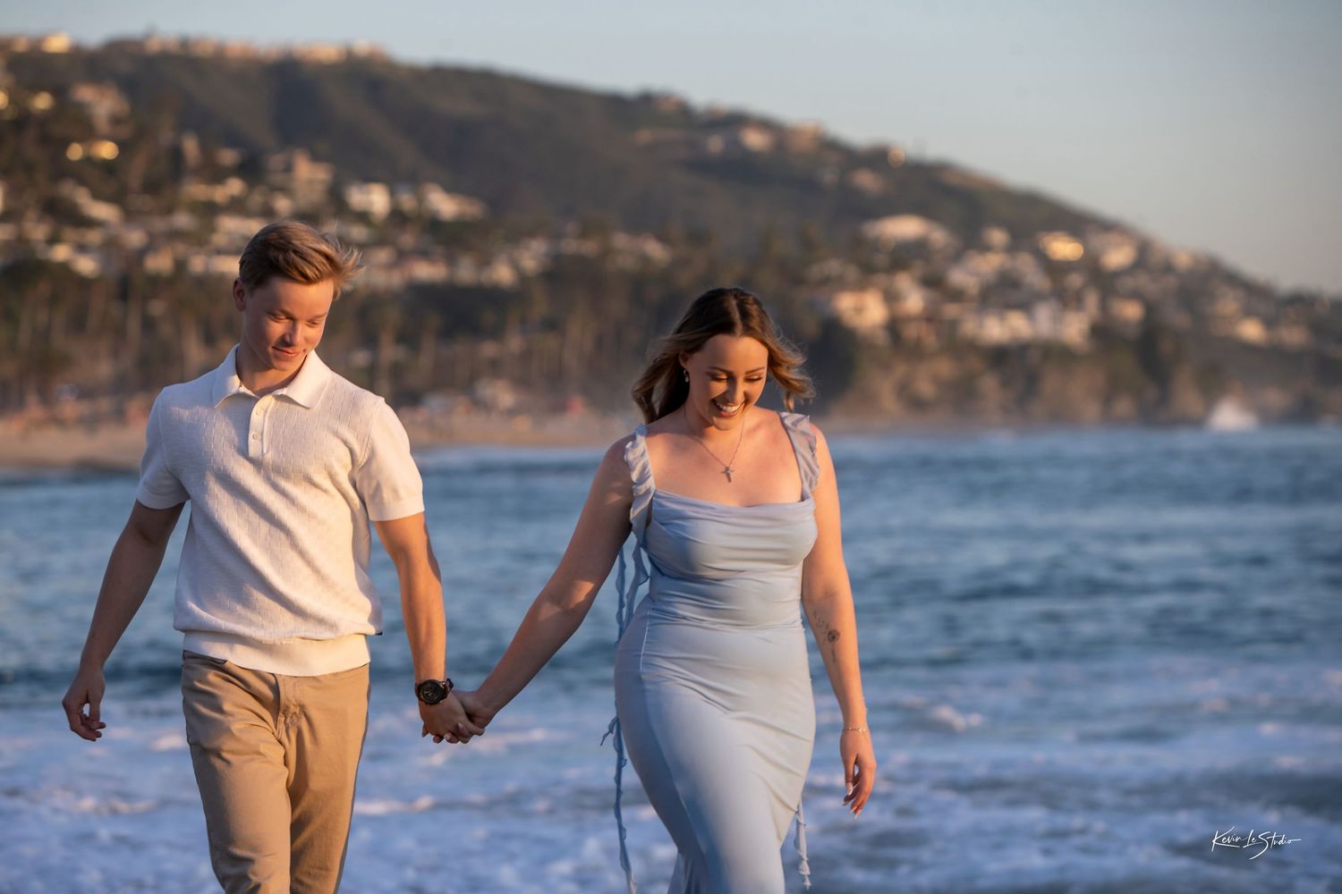 Young couple smiling and holding hands strolling along ocean shoreline at golden hour.