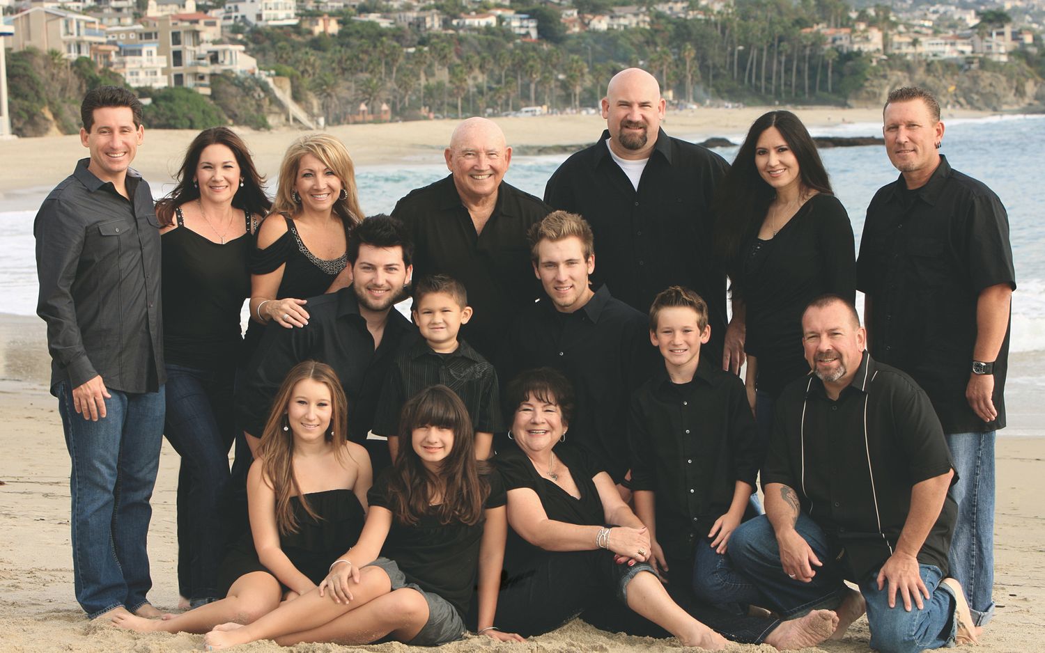 Large family group of fourteen posing together on a beach in coordinating black outfits.