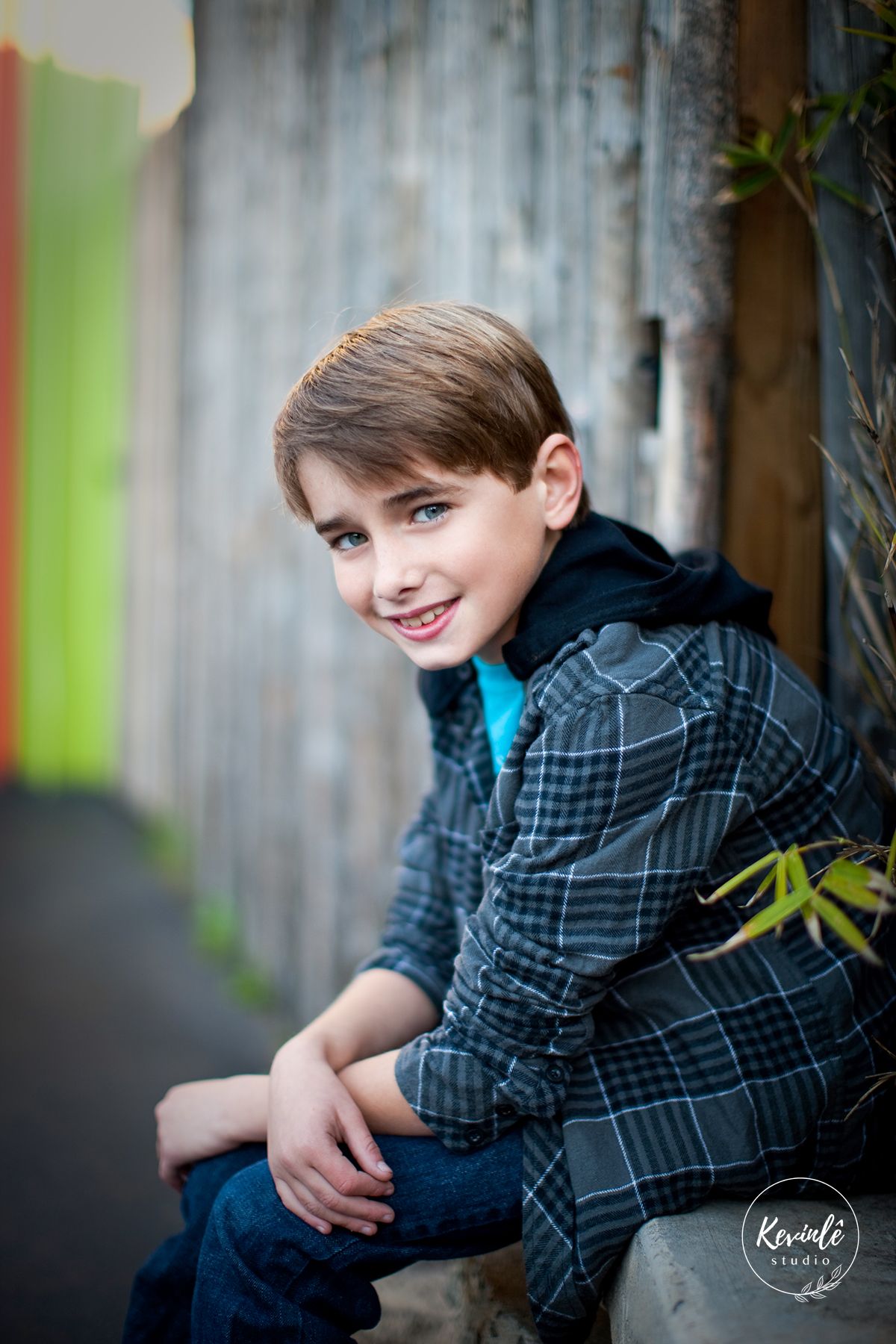 A smiling boy in a plaid jacket sits outdoors against a wooden fence in a professional portrait photo.