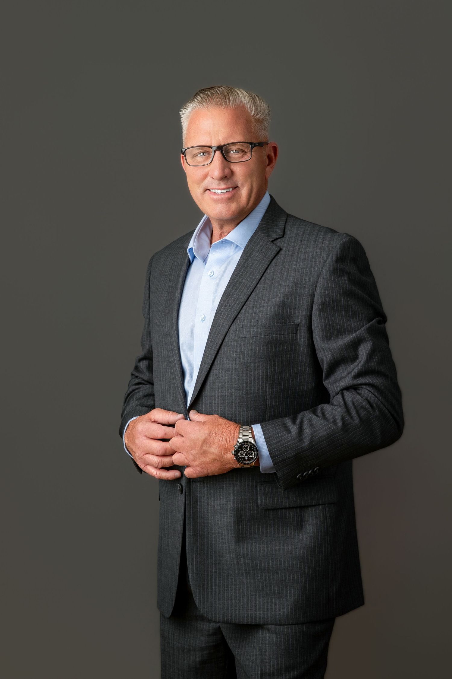 Professional man in gray pinstripe suit and glasses smiling confidently against dark background.