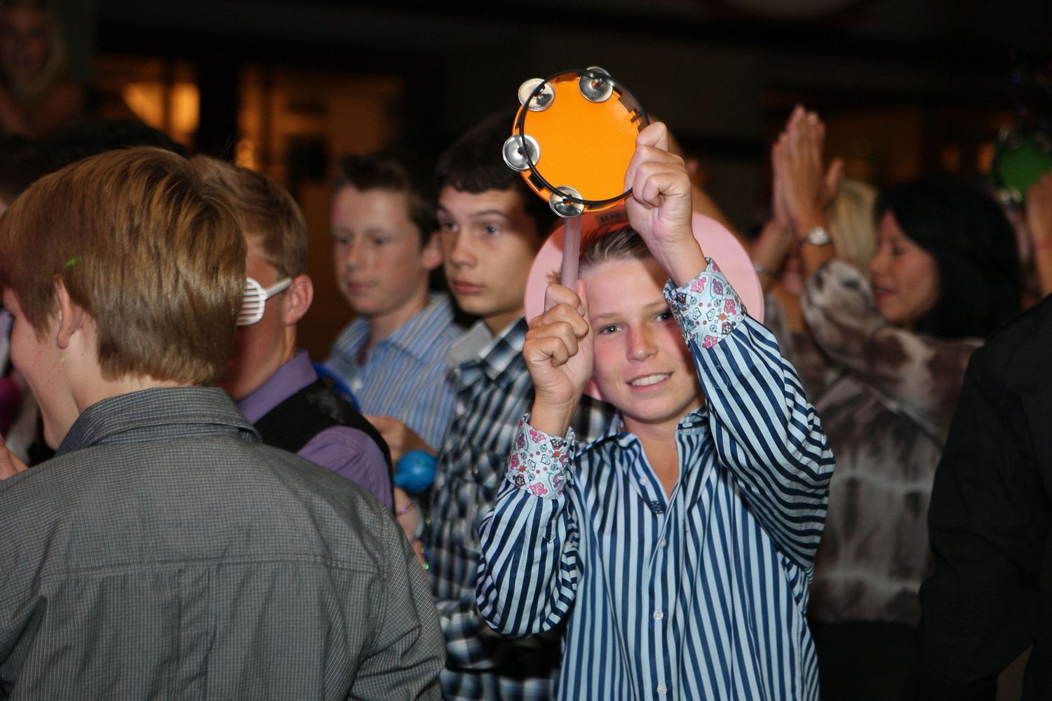 A smiling girl holds an orange tambourine above her head at an indoor party event with other children nearby.