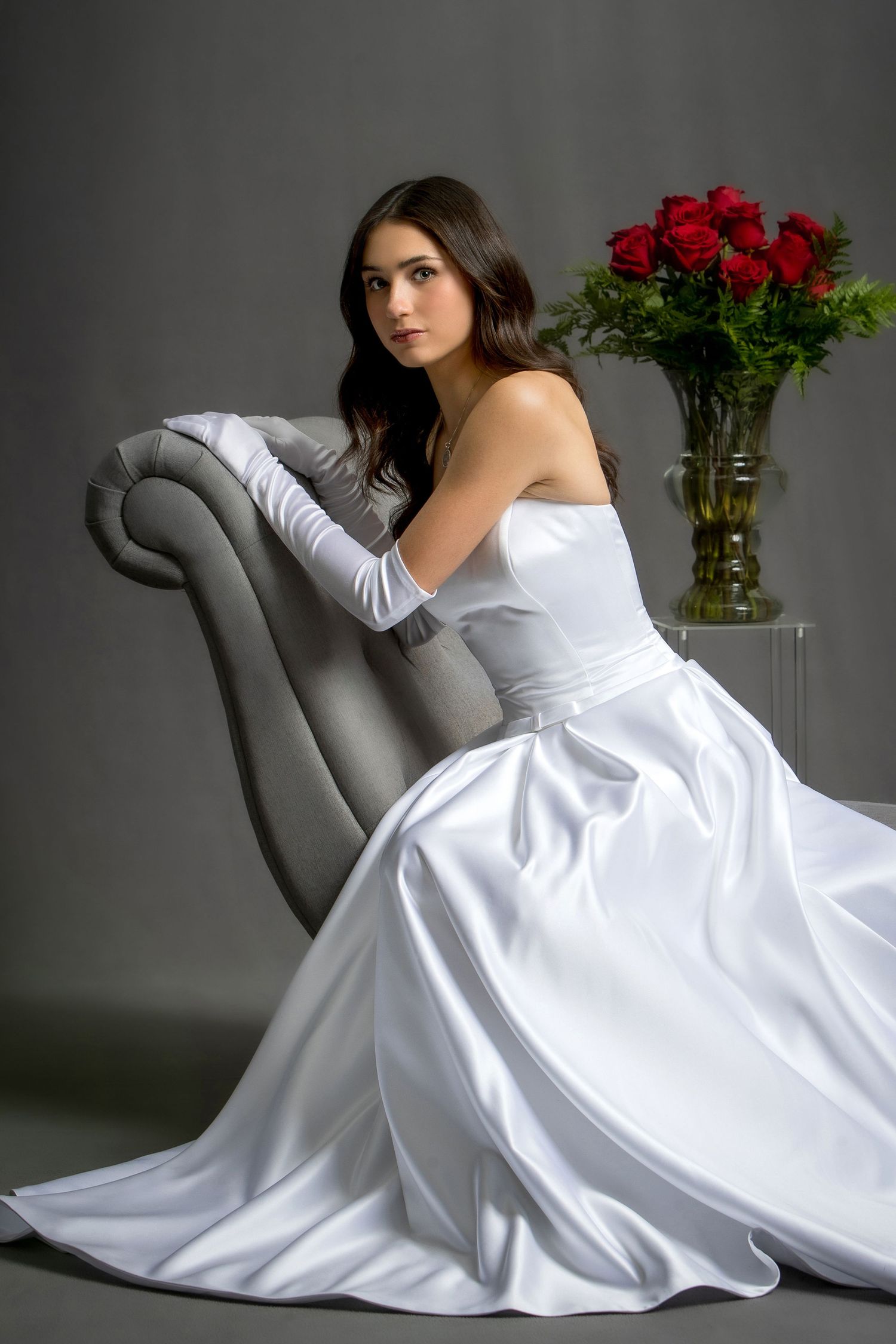 Elegant bride in white satin strapless gown and gloves seated on gray chair beside red roses bouquet.