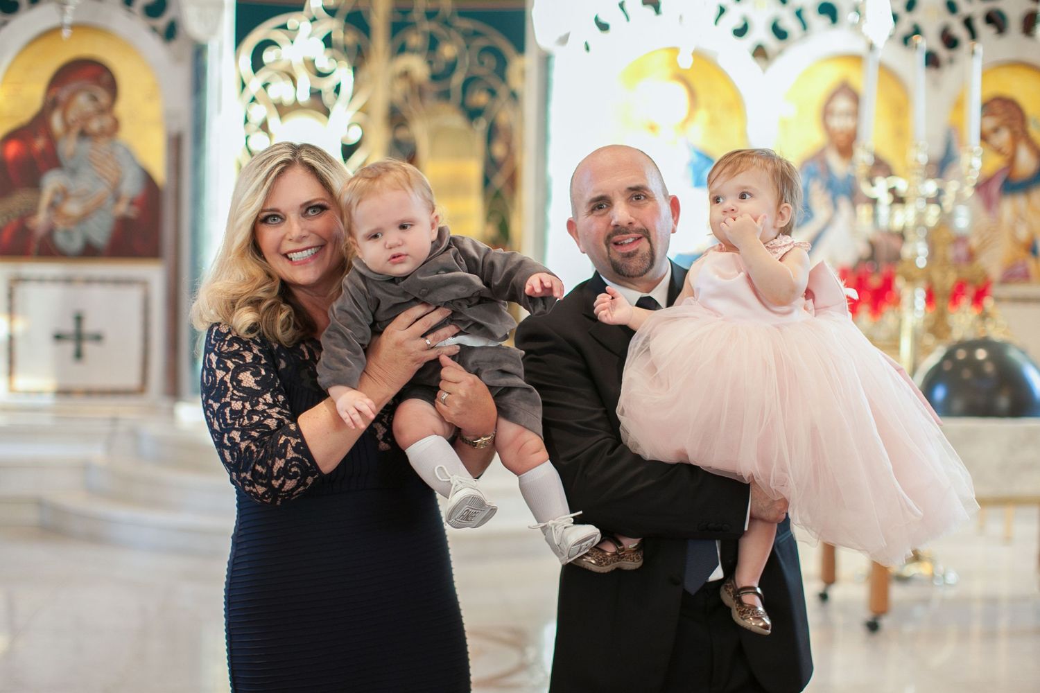 Family of four posing in an ornate Orthodox church, parents holding two toddlers dressed in formal attire.