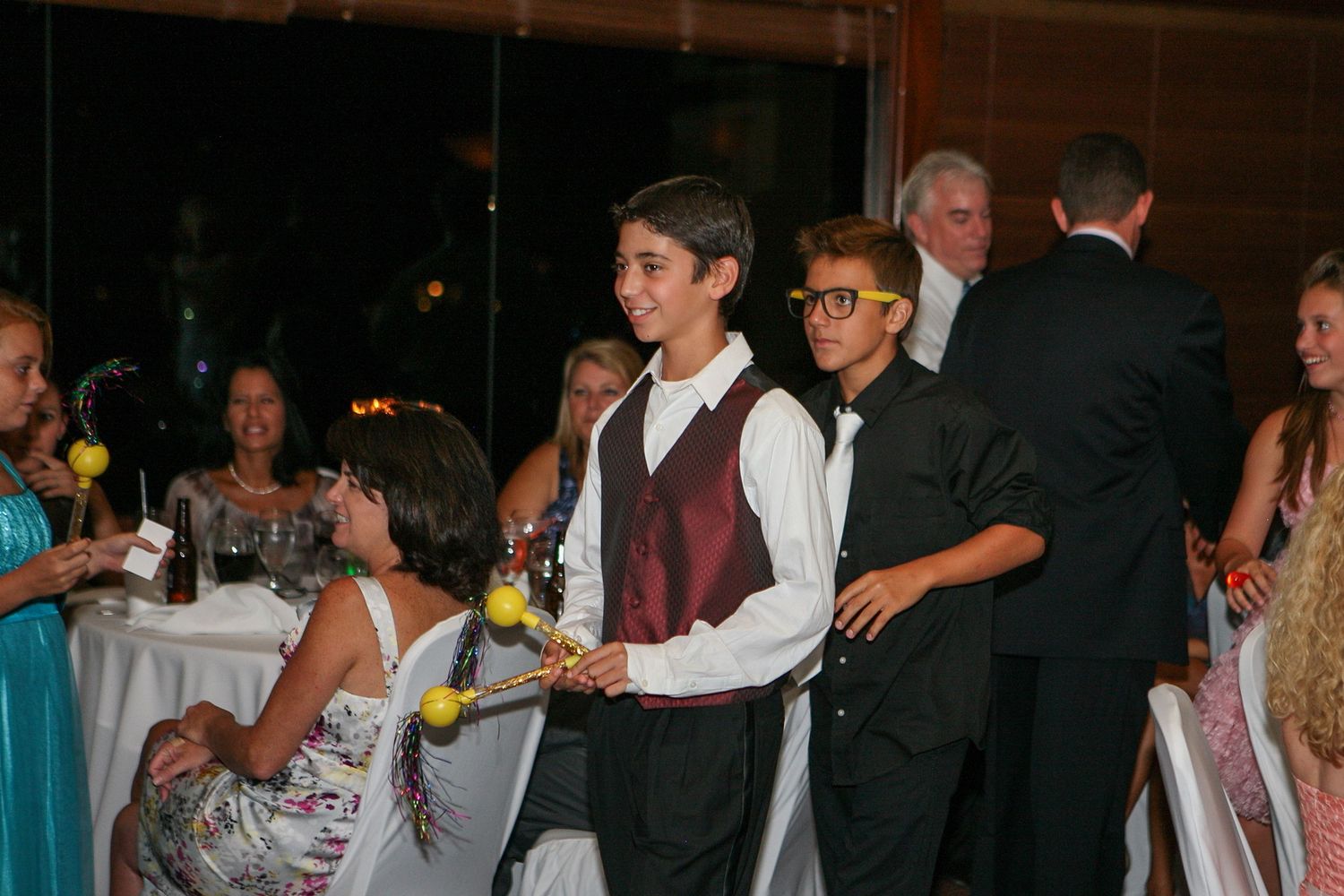Two boys in formal attire smile at a festive indoor event, surrounded by guests at a celebration.