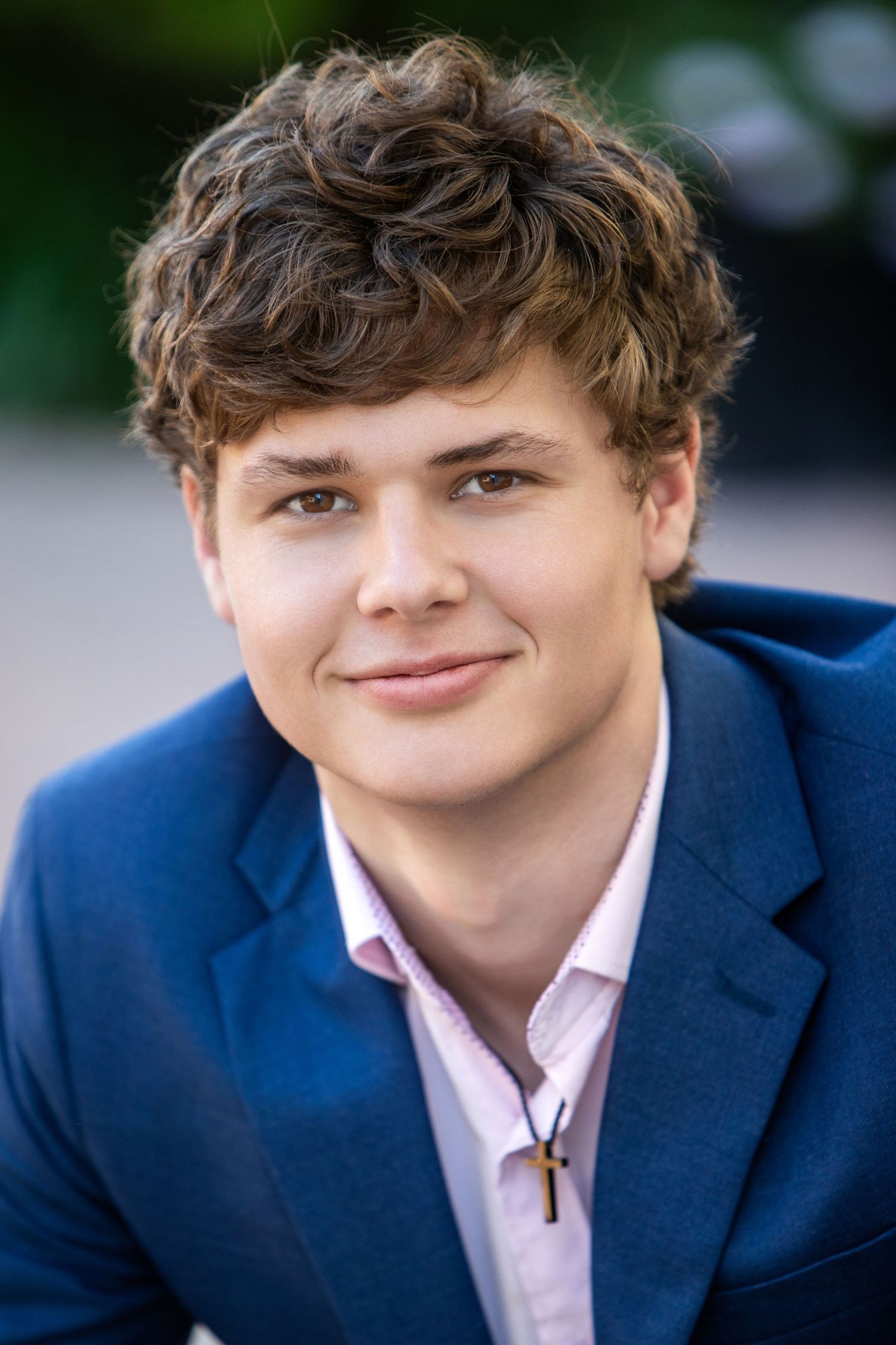 Young man with curly brown hair smiling in a blue blazer and pink shirt with a cross necklace.