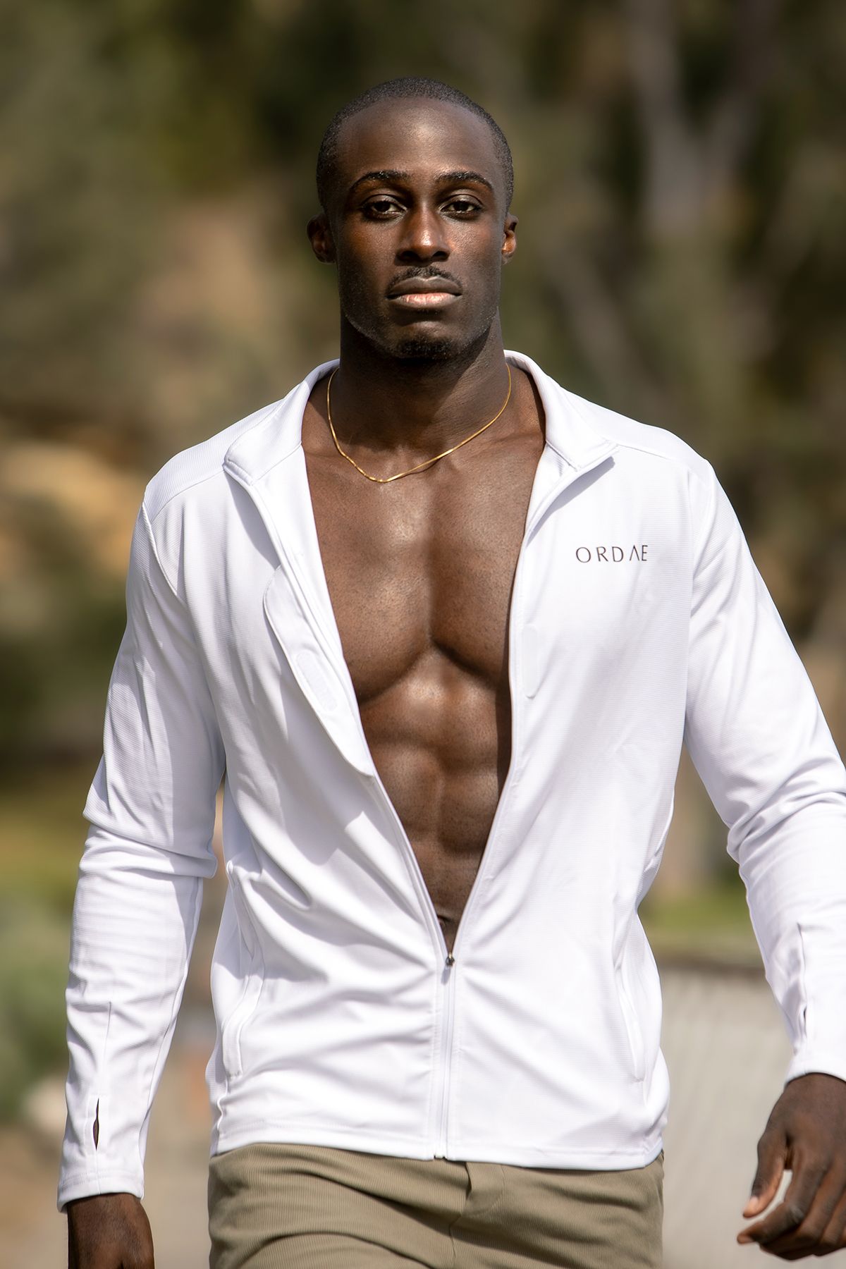 Athletic Black man in open white zip-up jacket walking outdoors with trees in background.