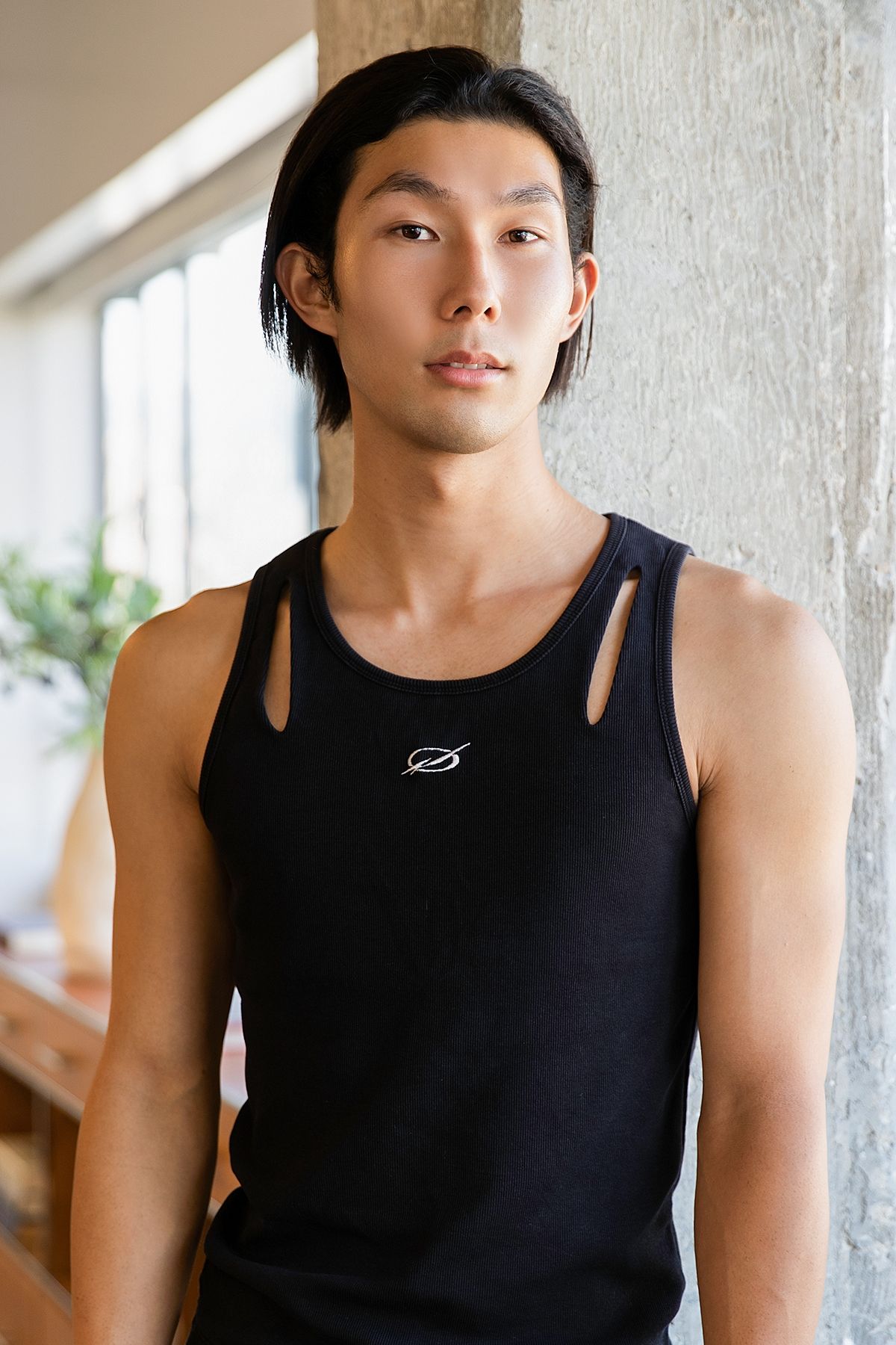 Young Asian man with black hair wearing a black sleeveless tank top, posing against a concrete wall.