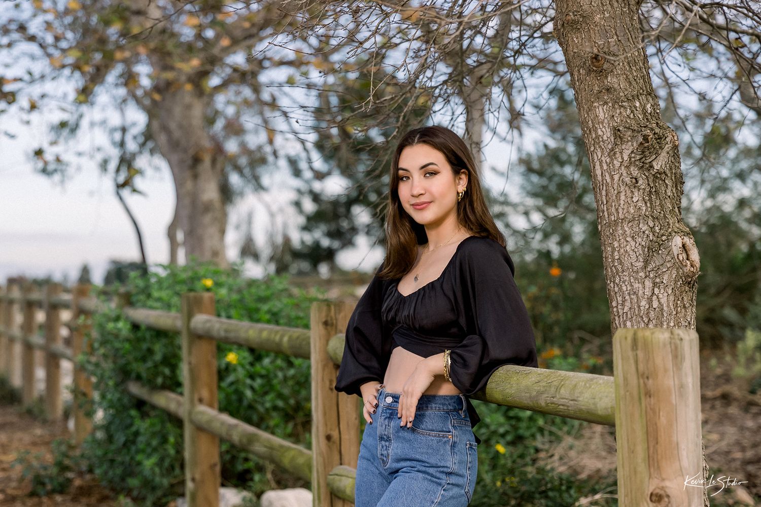 Young woman in black crop top and jeans poses by wooden fence surrounded by trees and greenery.