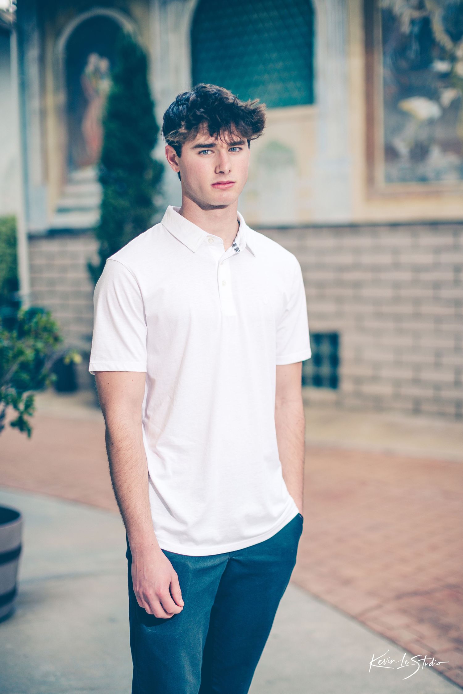 Young man in white polo shirt and teal pants standing outdoors in a European-style courtyard.