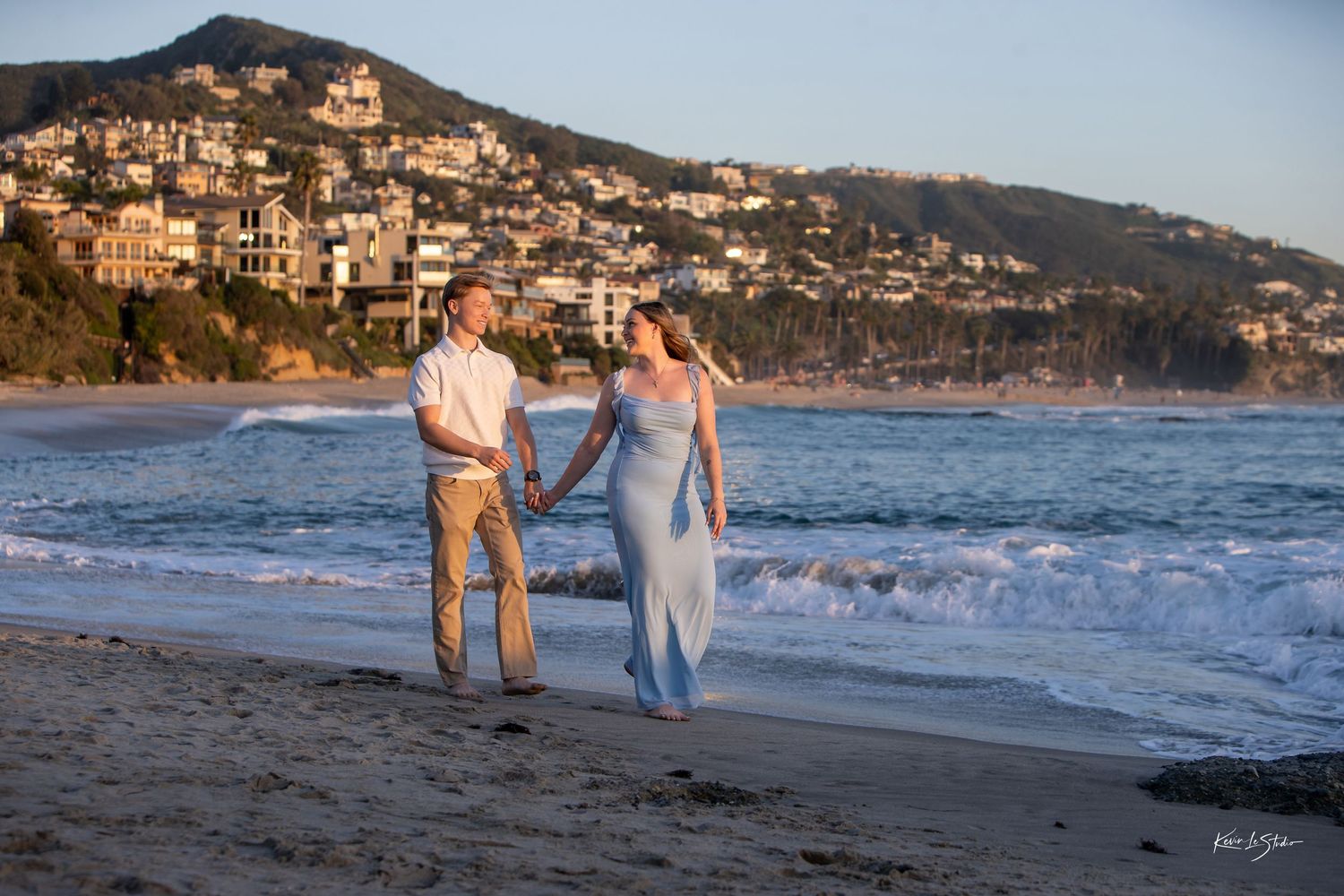 Couple holding hands walking on beach at sunset with Laguna Beach hillside homes in background.