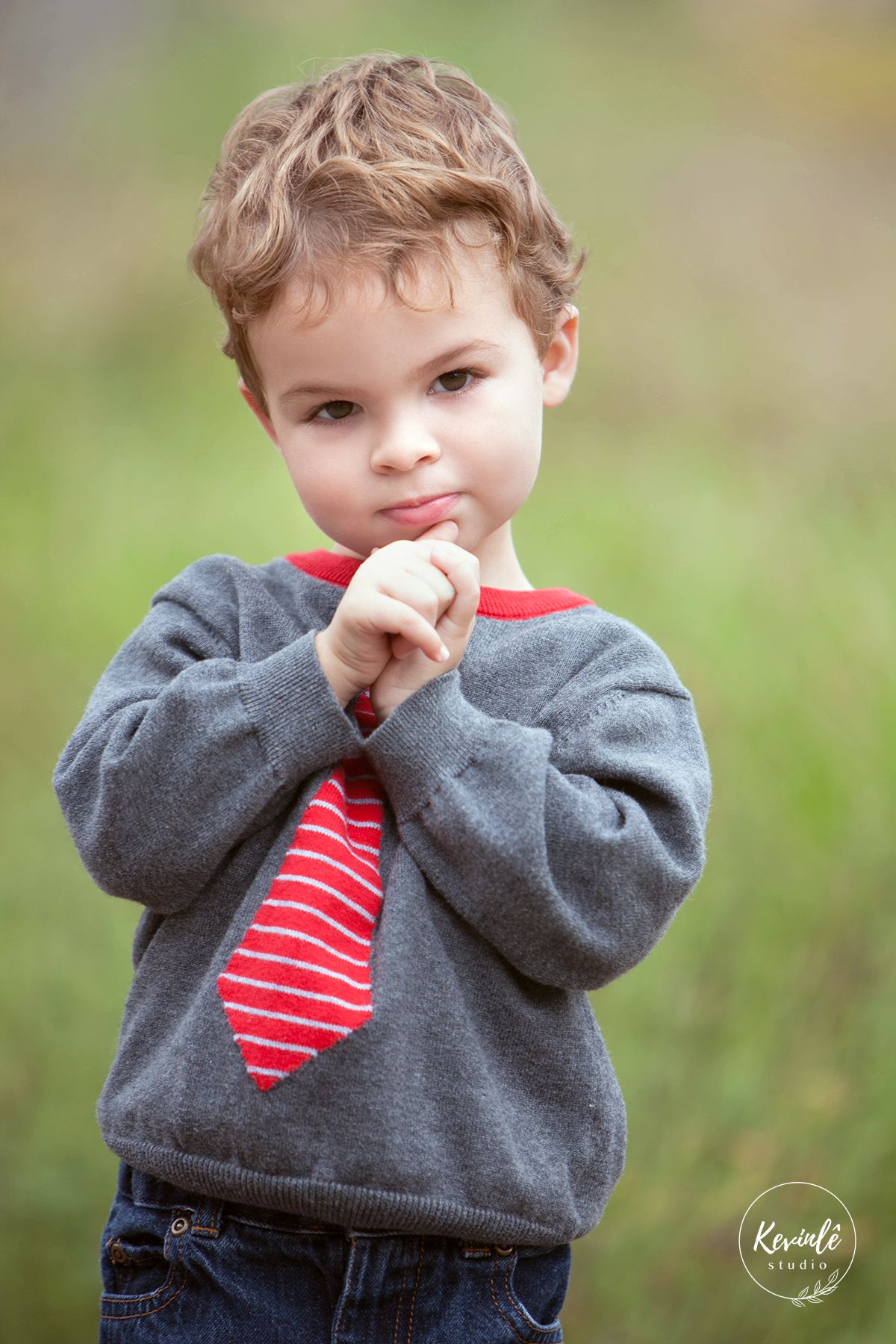 Cute toddler boy in a gray sweater with a red striped tie posing outdoors in a green field.
