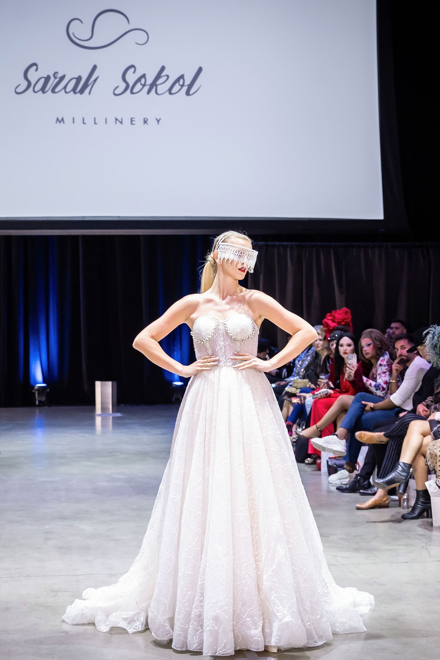 Blonde model in a Sarah Sokol Millinery headpiece and elegant blush ballgown poses confidently on a fashion runway.
