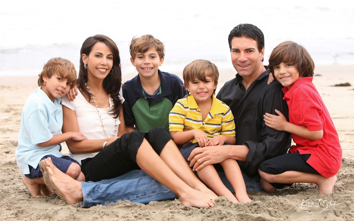 Happy family of six posing together on a sandy beach, smiling for a portrait on a cloudy day.