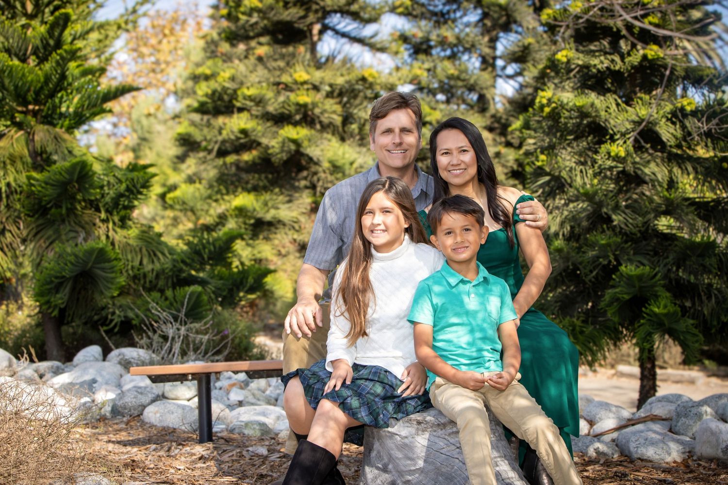 Happy family of four smiling together outdoors on rocks surrounded by lush green pine trees on a sunny day.