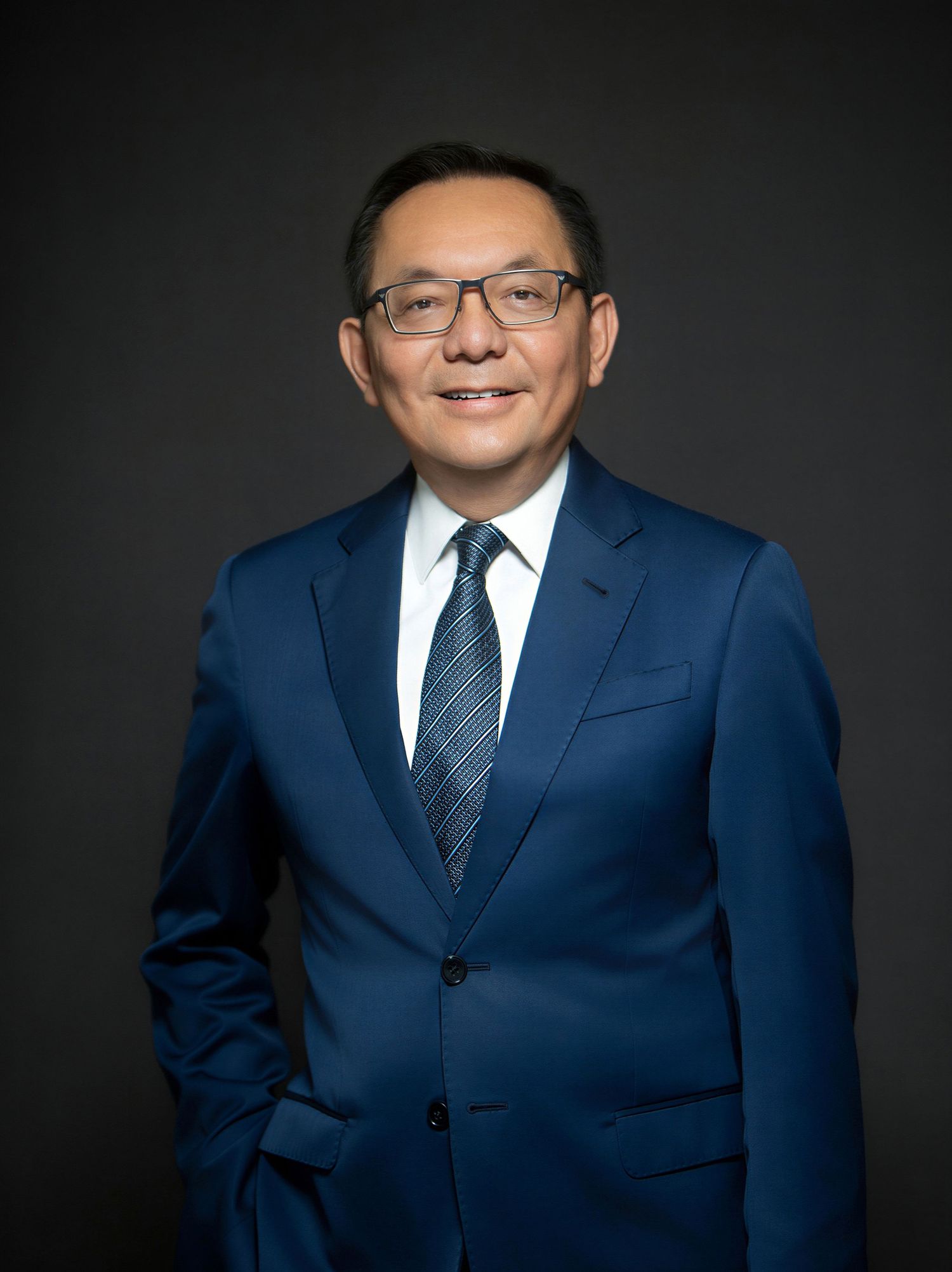 Professional headshot of a smiling man in a blue suit, white shirt, and striped tie against a dark background.