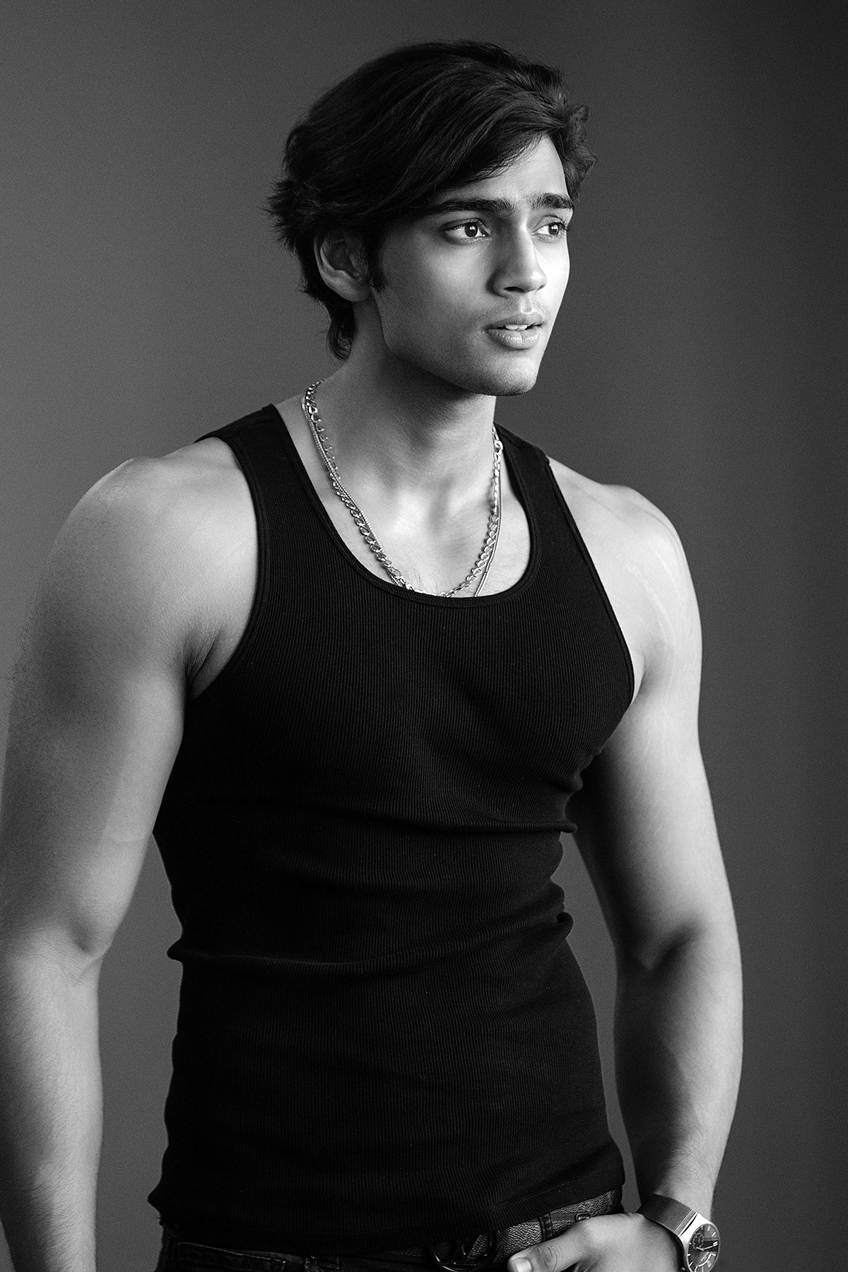 Young muscular man in black tank top and chain necklace poses in black and white studio portrait.