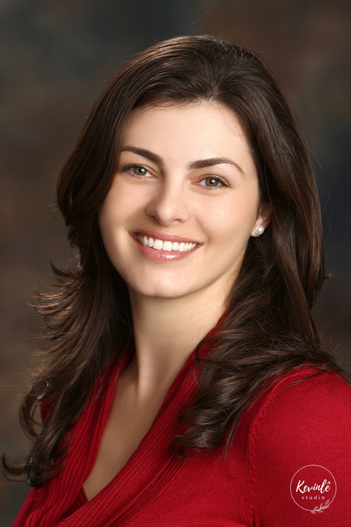Professional headshot of a young brunette woman with green eyes wearing a red top on a studio background.