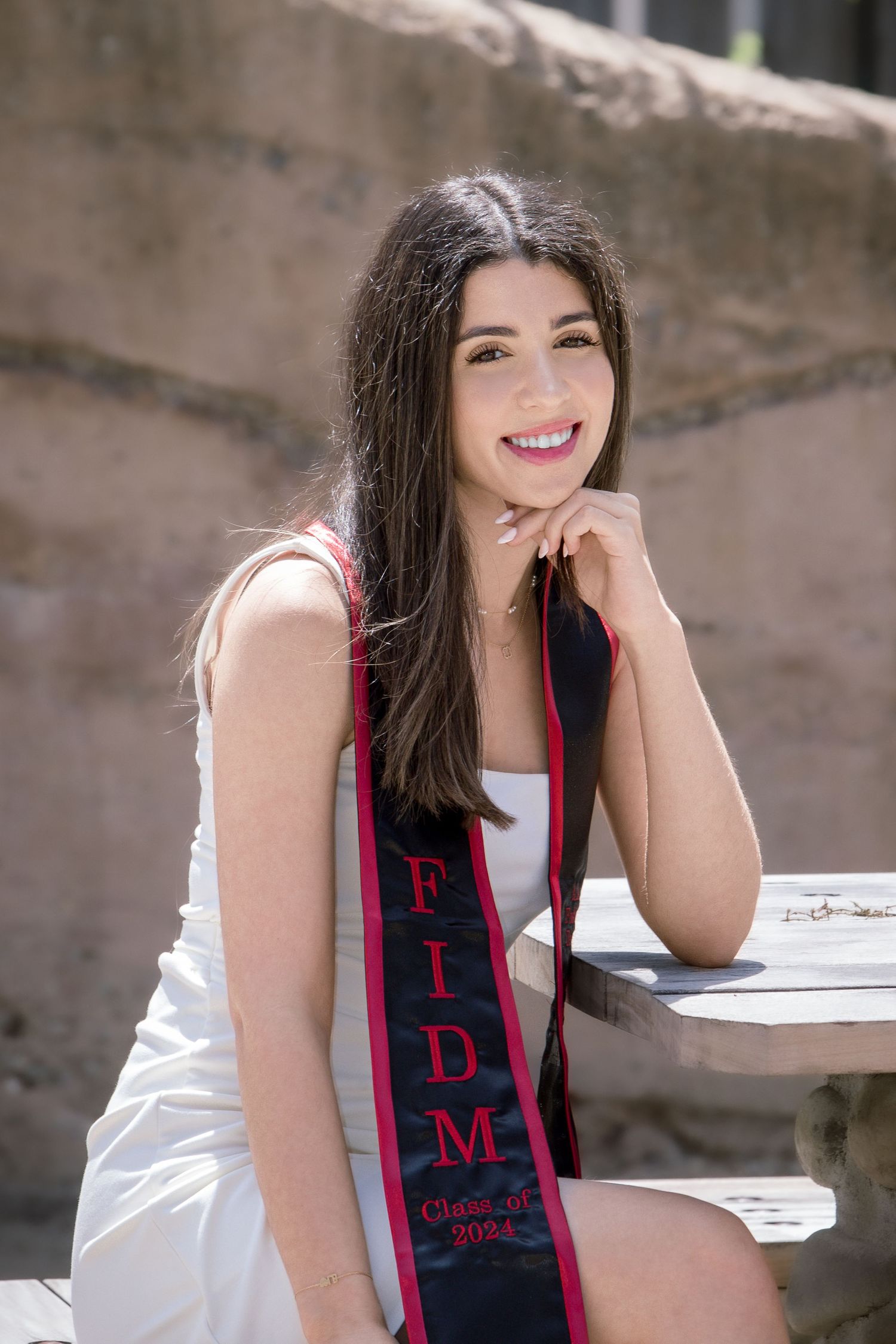 Young woman in white dress wearing FIDM graduation sash smiles outdoors in senior portrait.