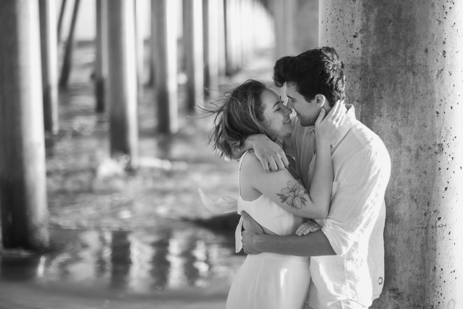 Black and white romantic photo of a couple about to kiss under a wooden pier by the water.