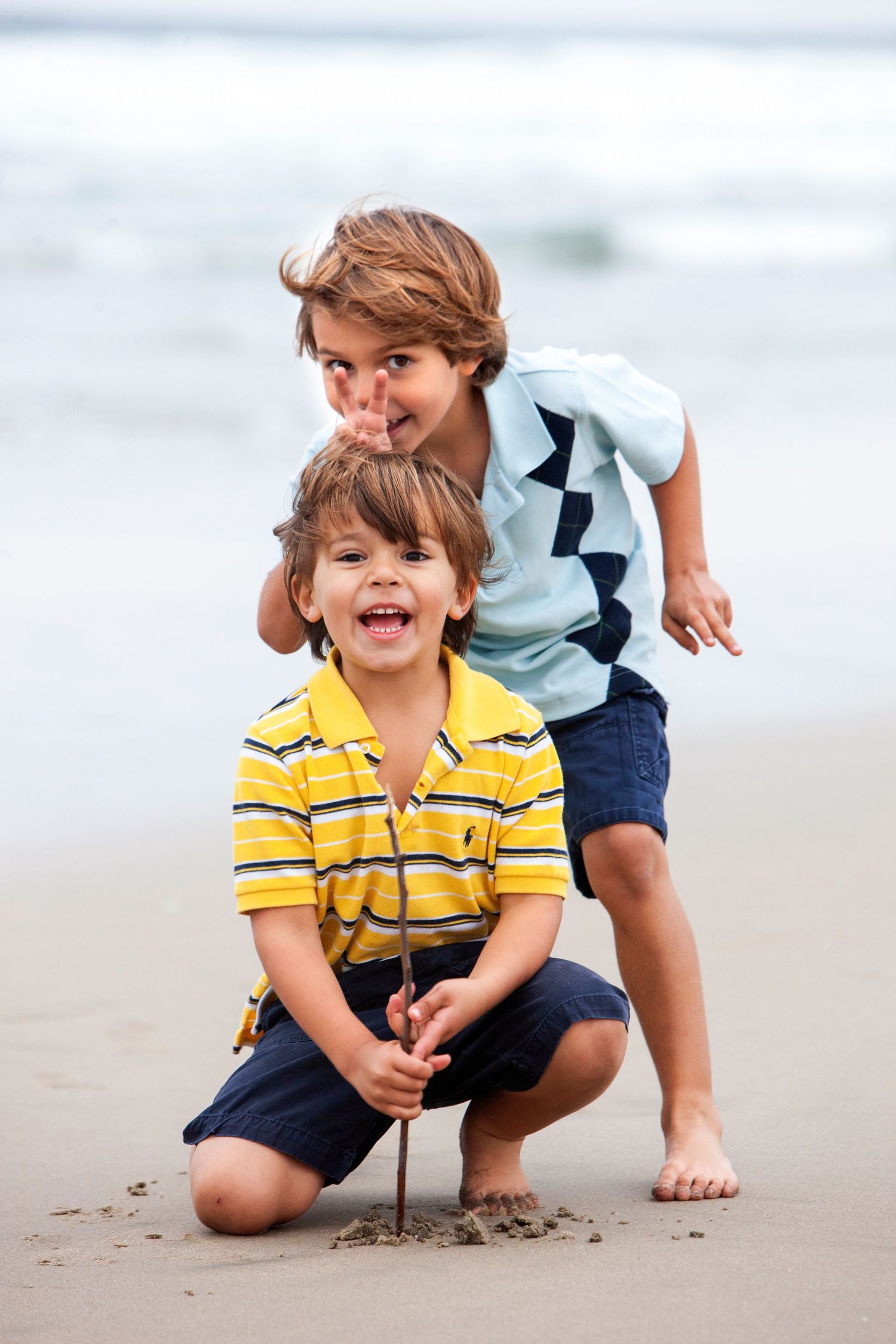 Two young boys playing on a sandy beach, one crouching with a stick while the other stands behind smiling.