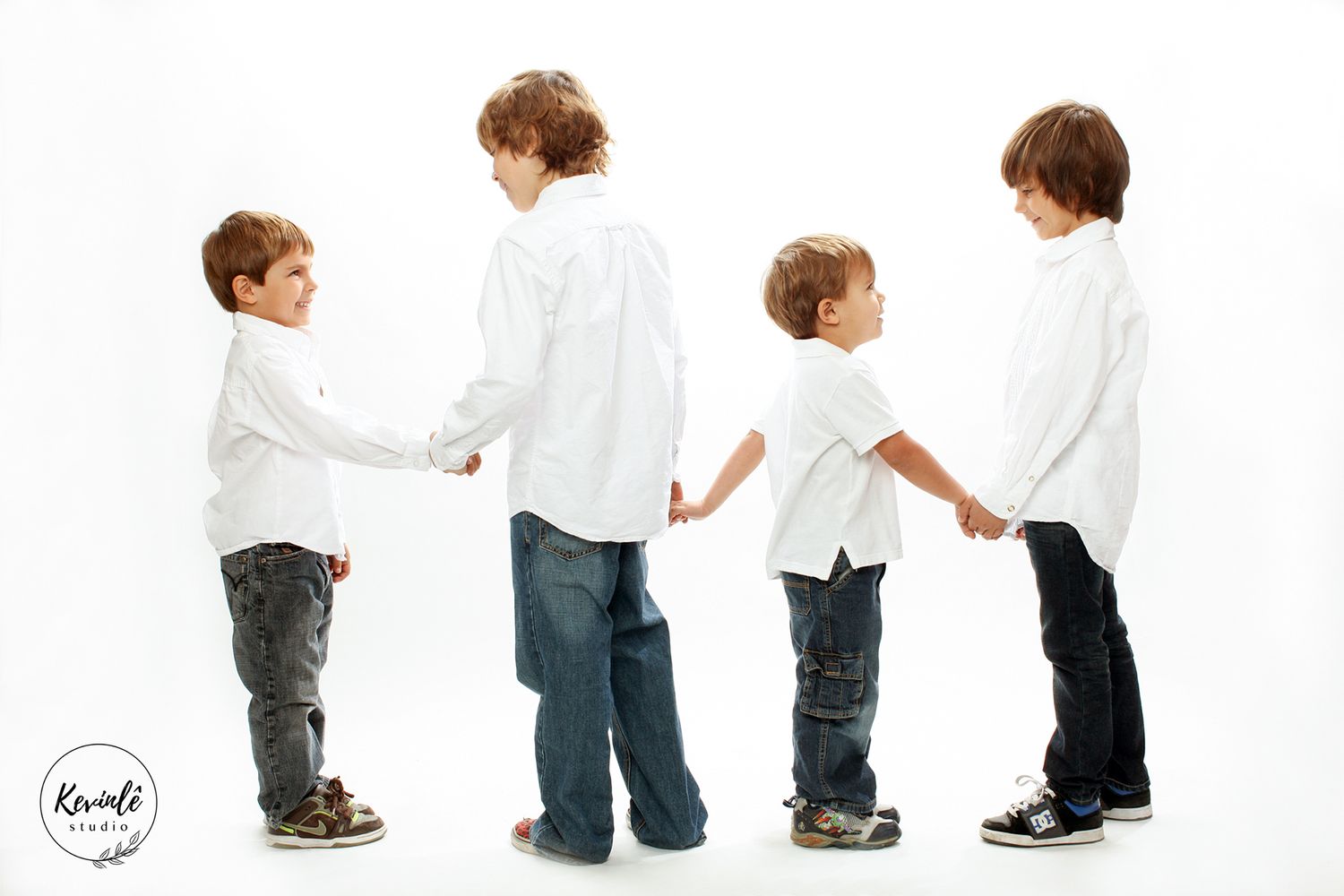 Four young boys in white shirts and jeans holding hands in a row against a white studio background.