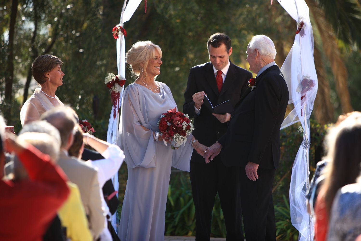 Outdoor wedding ceremony with bride in blue dress, groom in dark suit, and officiant under floral arch.