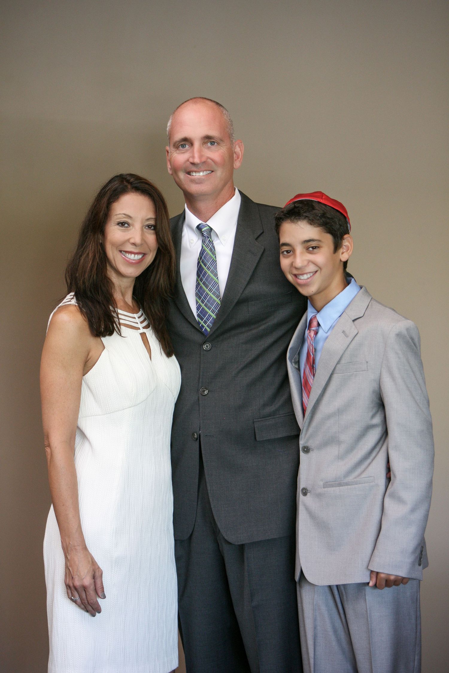 Family of three posing for a formal photo, woman in white dress, man in gray suit, teen boy in light suit with red kippah.