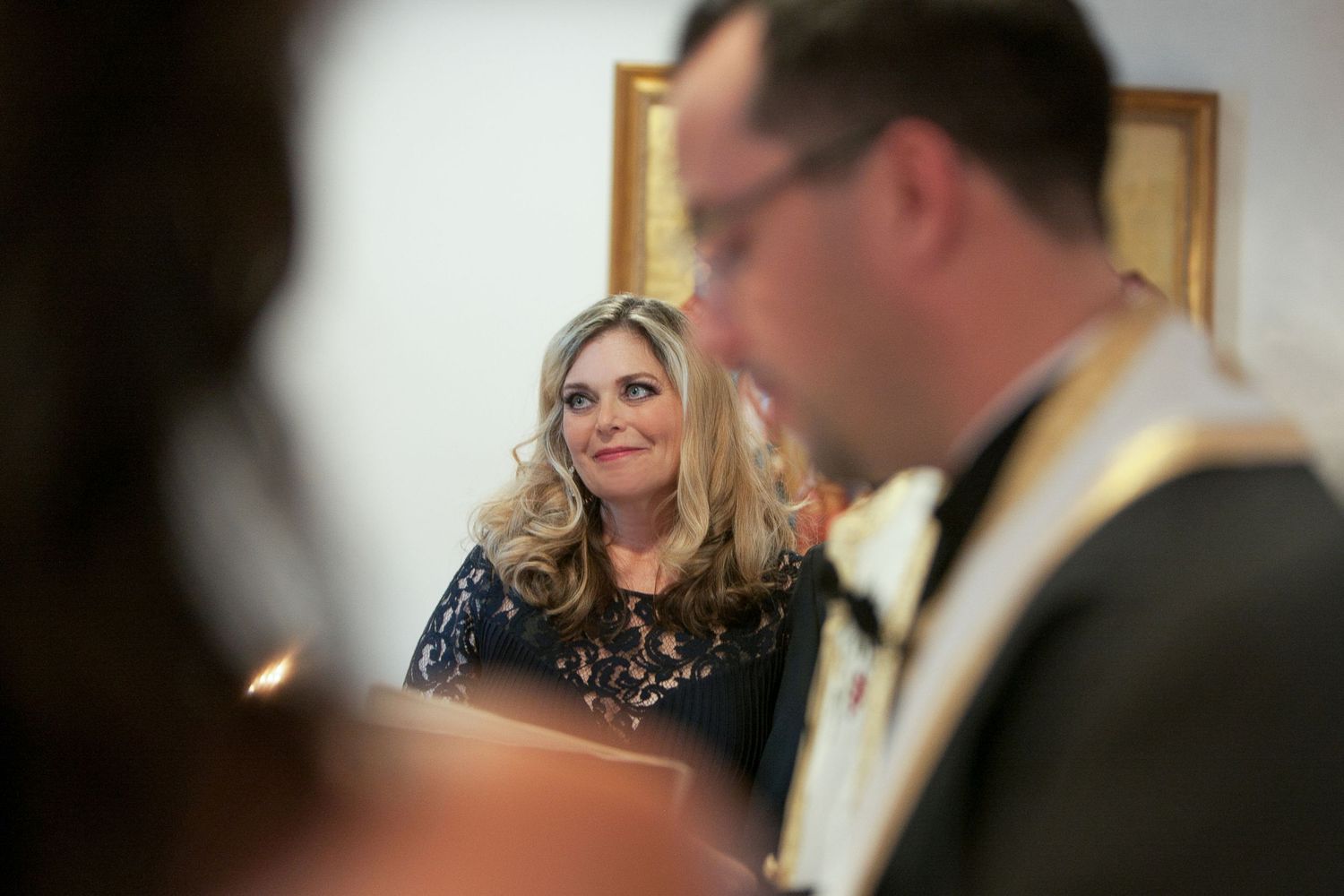 A woman in a navy lace dress smiles warmly during a formal ceremony, with blurred figures in the foreground.