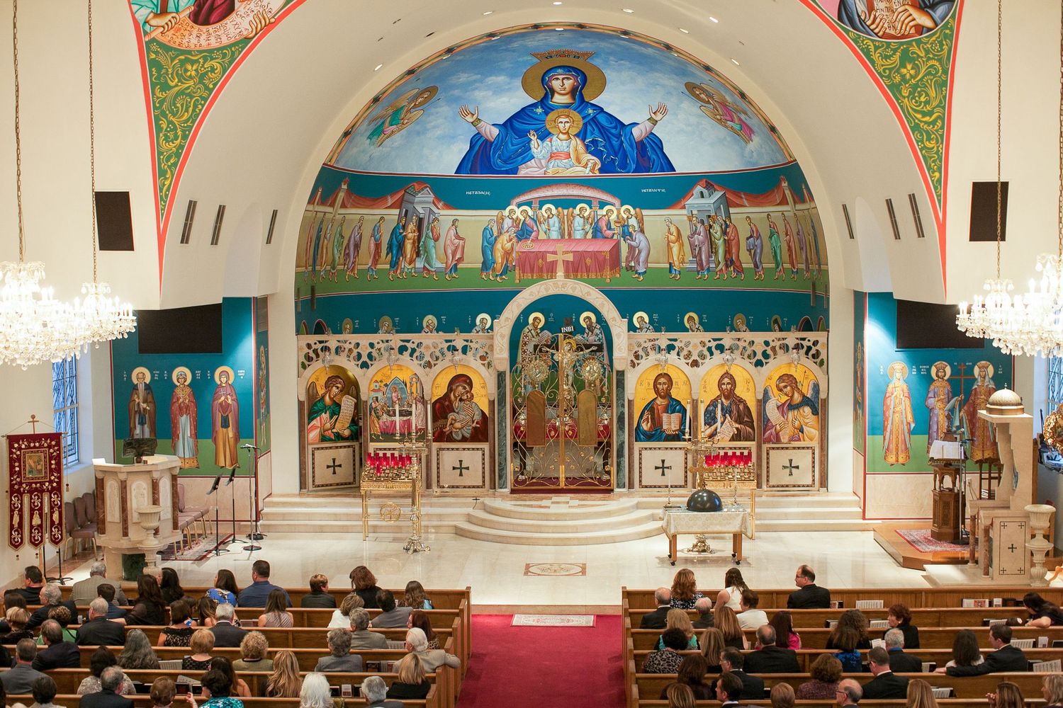Orthodox Christian church interior with ornate Byzantine murals, iconostasis, and congregation seated in wooden pews.