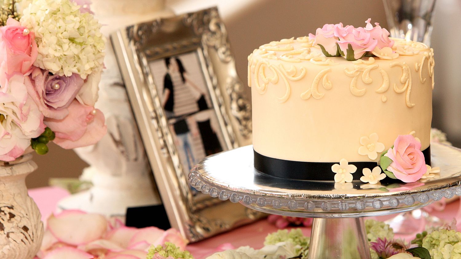Elegant white wedding cake with pink roses on a silver stand, surrounded by floral arrangements and vintage frames.