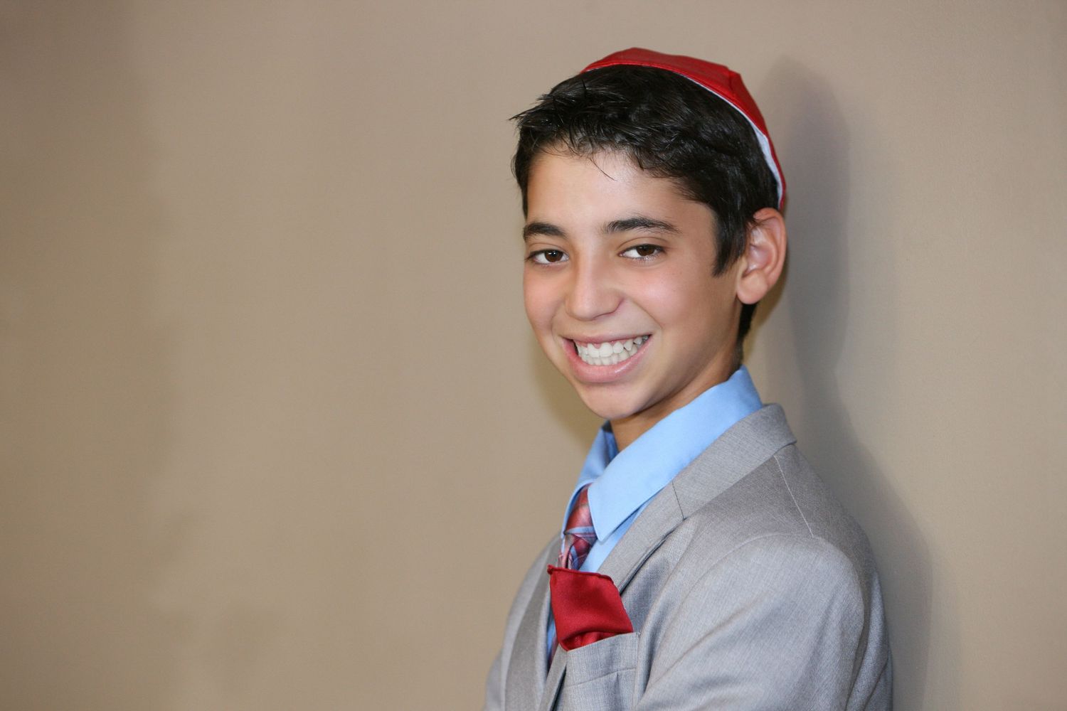 Smiling young boy in gray suit, red bow tie, and red kippah posing for a Bar Mitzvah portrait.