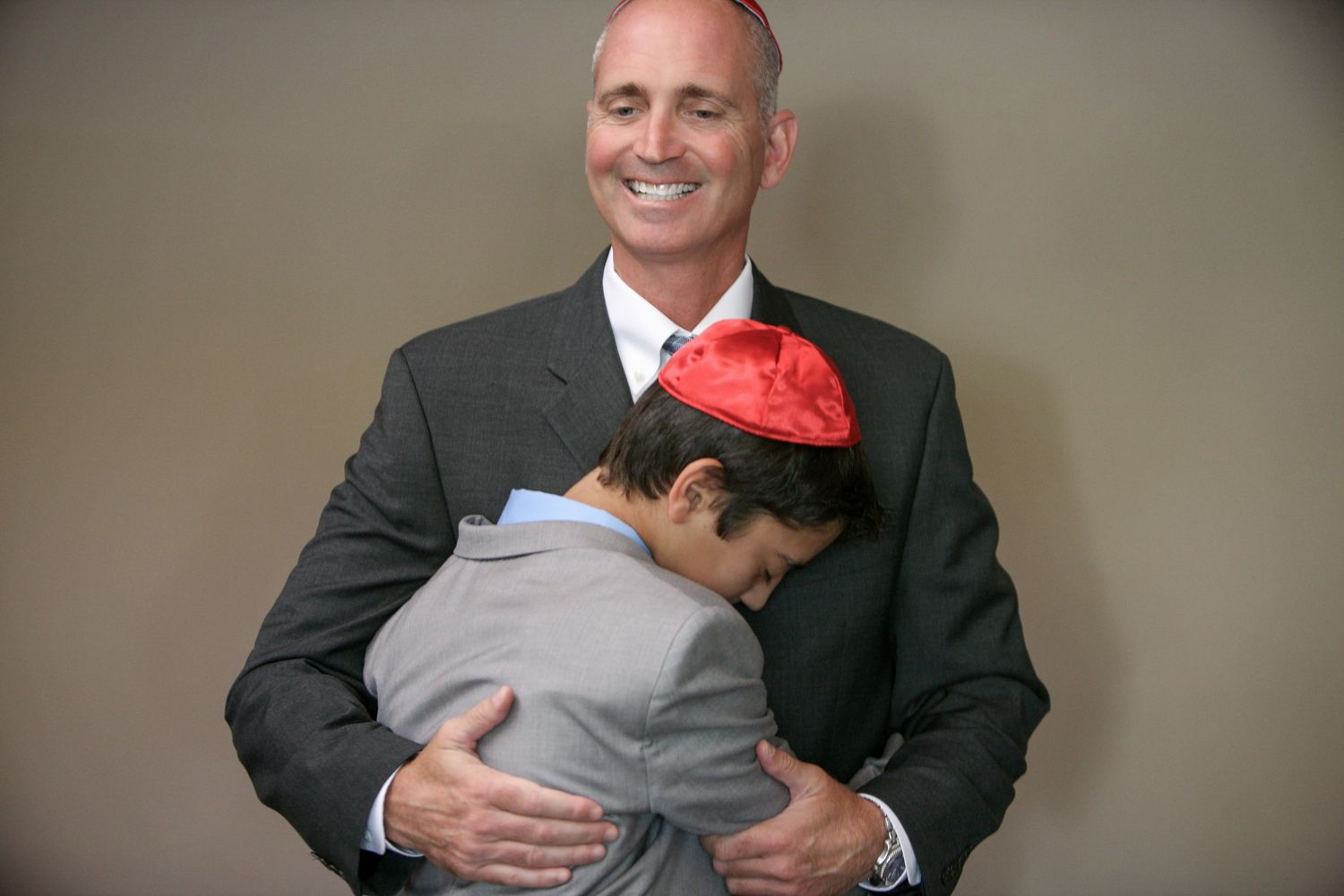 A smiling man in a suit hugs a boy wearing a red kippah during a Jewish celebration.