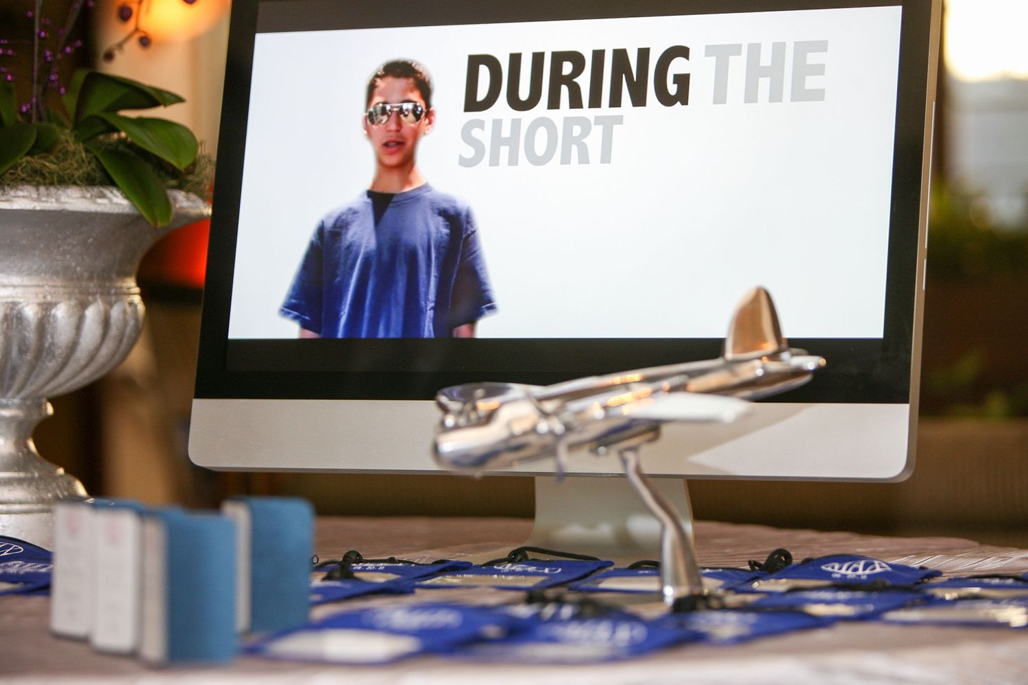 A desktop monitor displaying a man with text 'During The Short,' beside a silver model airplane on a blue tablecloth.