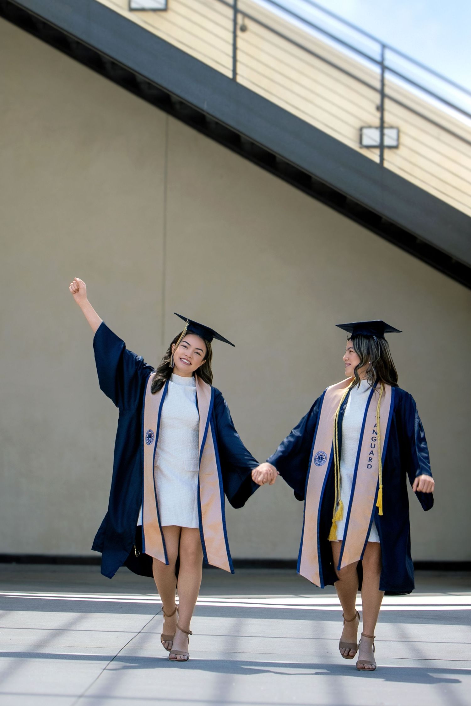 Two happy graduates in navy cap and gown holding hands and celebrating their graduation day outdoors.
